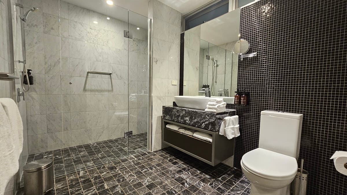 To the right is a toilet bowl positioned against the tiled wall. A tissue dispenser and a liquid soap dispenser are on both sides. To the left is a glass door securing the shower area. Shower and hand grabs are mounted to the wall, along with toiletries. A rubbish bin and a white towel are placed nearby. Adjacent is a lavatory with a hand basin and folded white towels on top. A towel organizer is placed underneath. The floor has printed black tiles, and the ceiling is white.