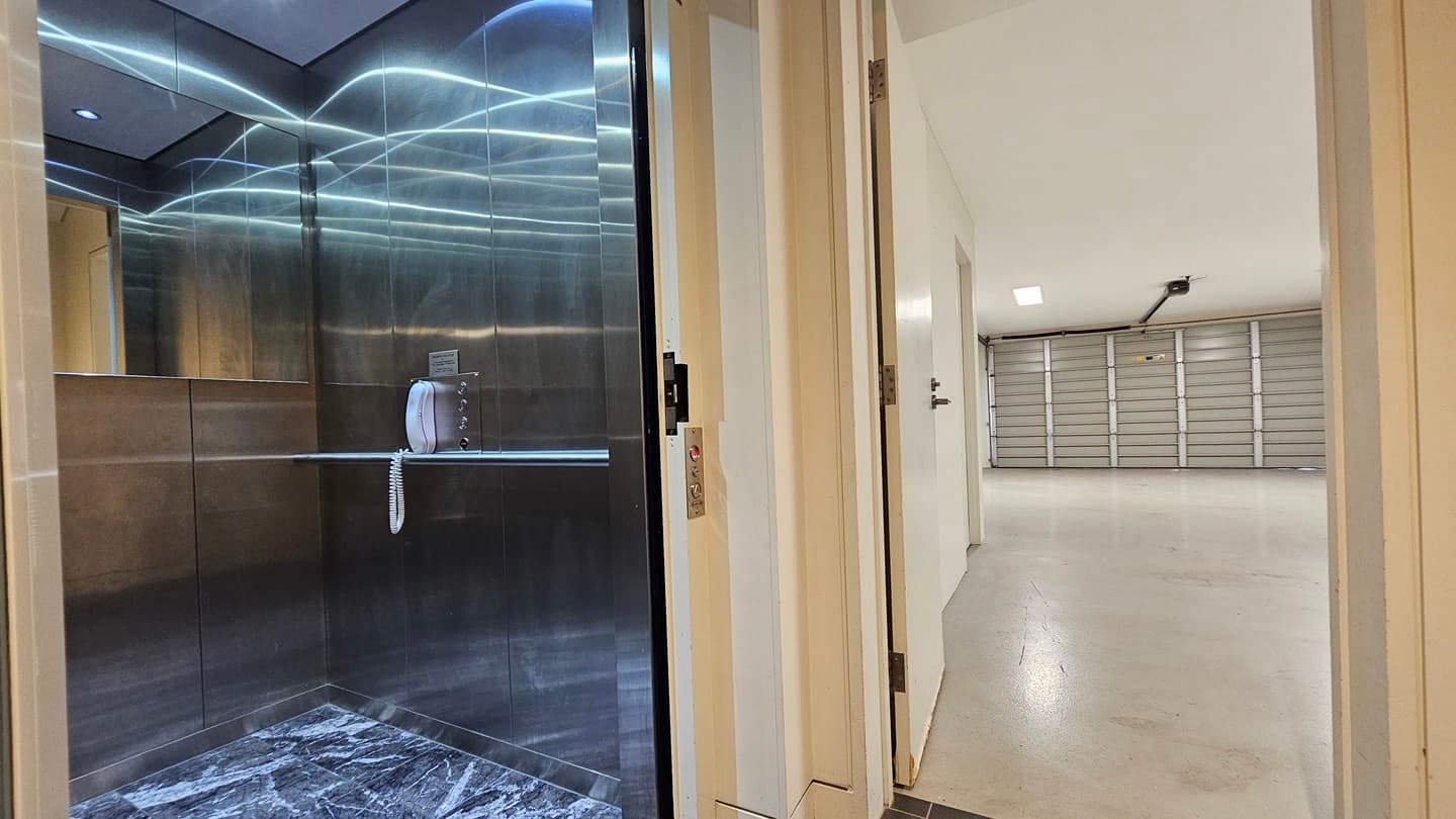 To the left is a lift equipped with mirrors, an emergency phone, and black printed floor tiles. To the right, a manual wooden door leads to the garage, an open space with white walls, white tiles, and a partition wall.