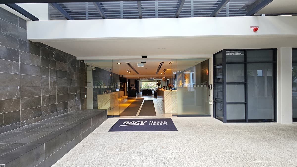 In the centre stands an automatic glass sliding door. A grey mat displaying the facility's name lies before the entrance. The left wall features dark grey tiles extending inside. On the right, a glass wall. Inside, a marble-designed reception desk with large paintings behind it. Opposite, another marble desk holds brochures and a flower vase. Nearby, a long cushioned bench chair for seating. The floor combines tiles and grey matting, while the ceiling, adorned with light fixtures, is white.
