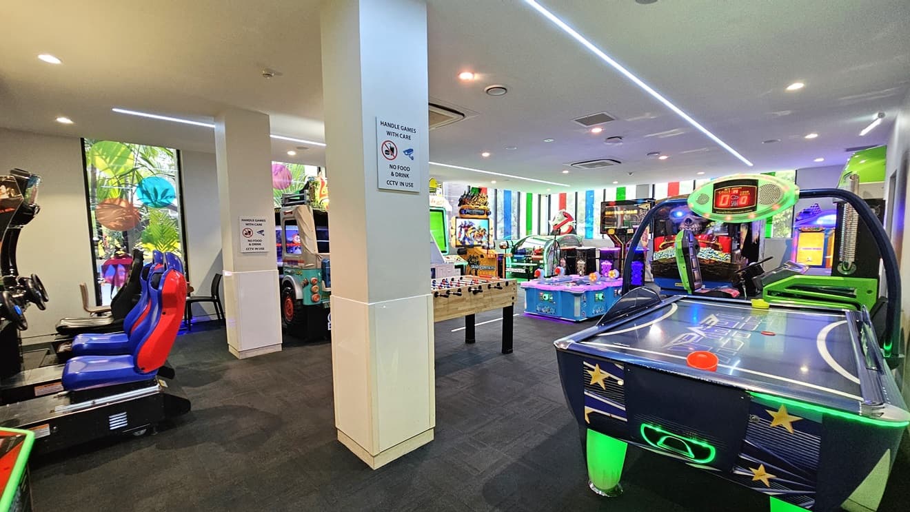 Outside, an outward-opening glass door with a hand sanitizer nearby. Inside, a black mat with the facility's name, a rubbish bin, and a large game poster near the entrance. Grey carpet covers the floor, while the ceiling and walls are white. A colorful poster isa affixed to the right wall. Game machines are visible indoors.