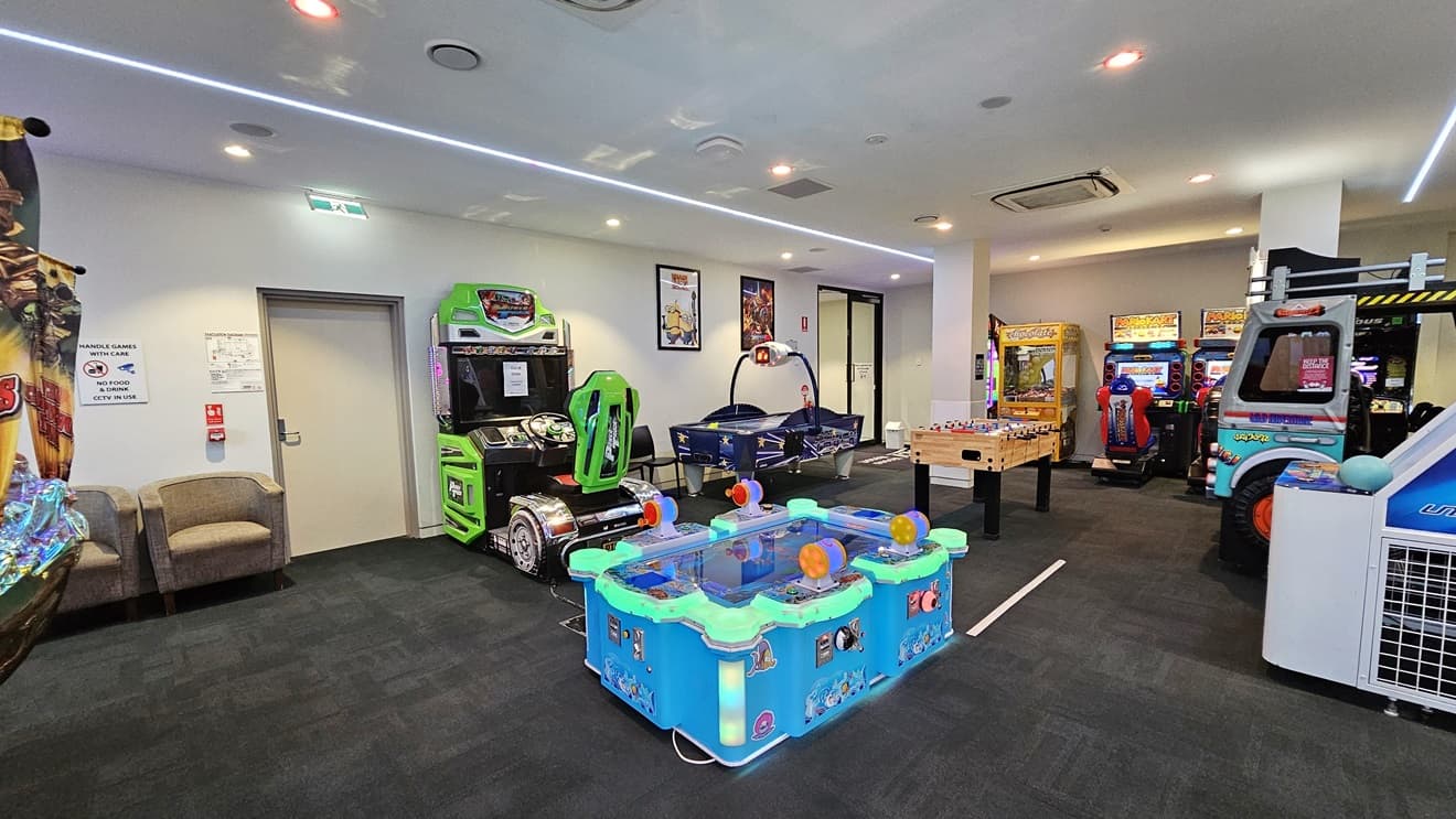 In the middle are electronic arcade games, a football table, and driving machines. A large riding machine is also on the right. Two cushioned grey chairs are positioned against the wall near the door on the left. The floor is carpeted in grey, and the walls have posters and wall decorations, including reminder posters. The ceiling is white with long fluorescent lights.