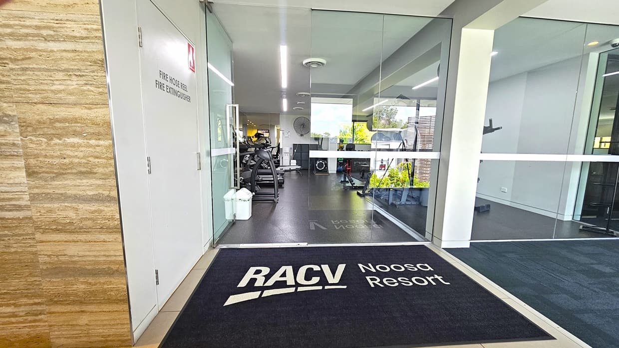 In the centre, there's a manual outward-opening glass door with a black mat displaying the facility's name. On the left wall, there's a fire hose reel and a fire extinguisher. The exterior wall has a timber design. Inside, the gym floor is covered with black rubber tiles. Gym equipment is arranged in the middle, and a white rubbish bin is placed near the entrance.