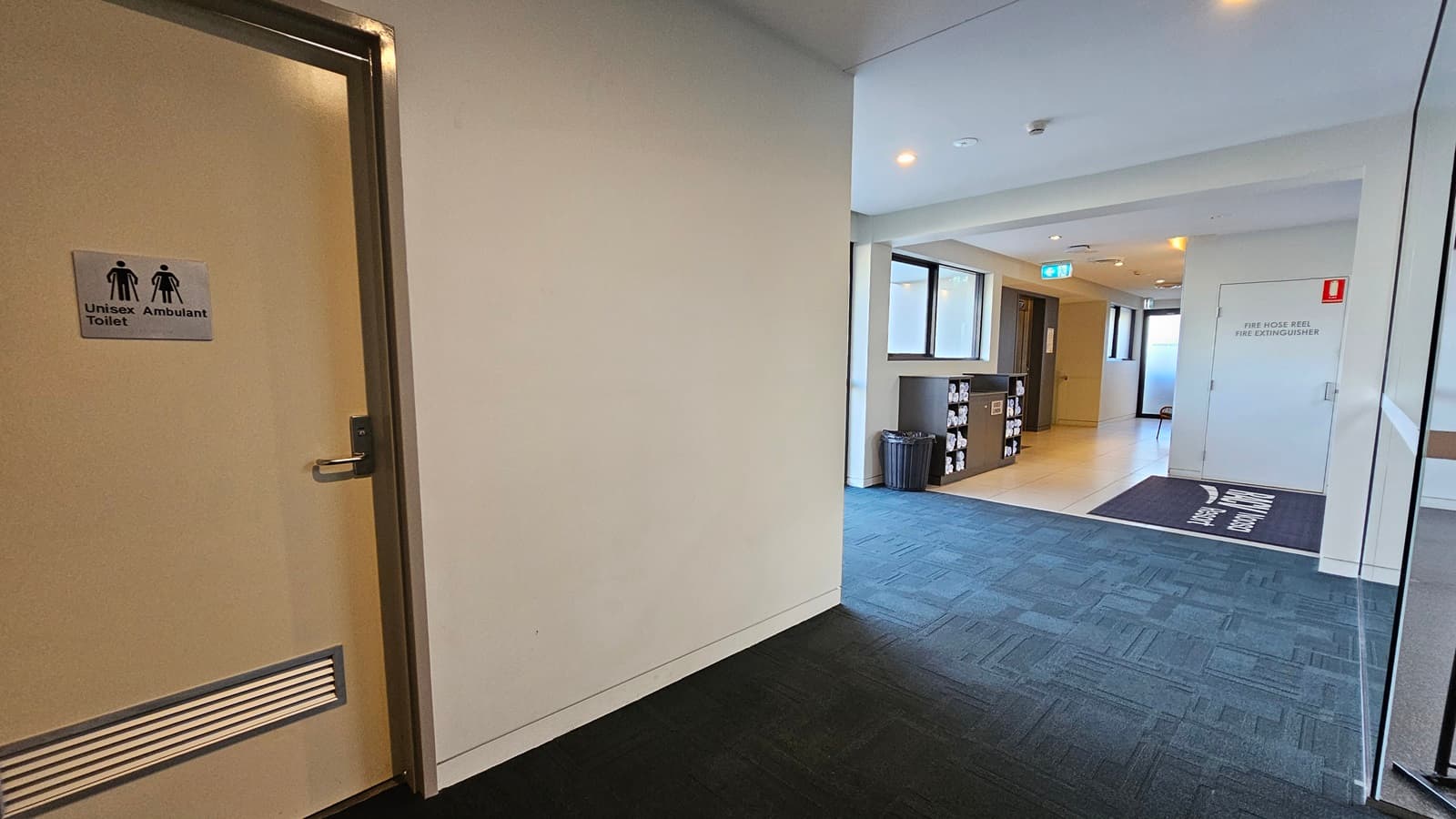 To the left is a Unisex Ambulant Toilet. To the right is a glass door that opens outward. A black matting bearing the facility's name is positioned on the door near the entrance. Further to the left is a display rack with rolled white towels against a glass window. A black rubbish bin is beside it. The floor transitions from grey to black rubber tiles inside.