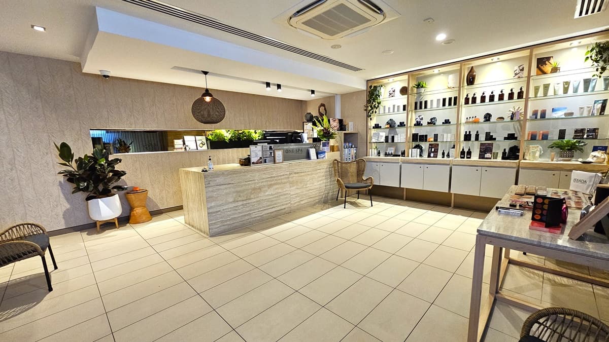 To the right is a timber wall with signage pointing to the Spa and Gym. Two rattan chairs and a small round table are positioned nearby. An indoor plant stands near the inward-opening glass door entrance. The floor has white tiles, and the ceiling is white with LED lights.