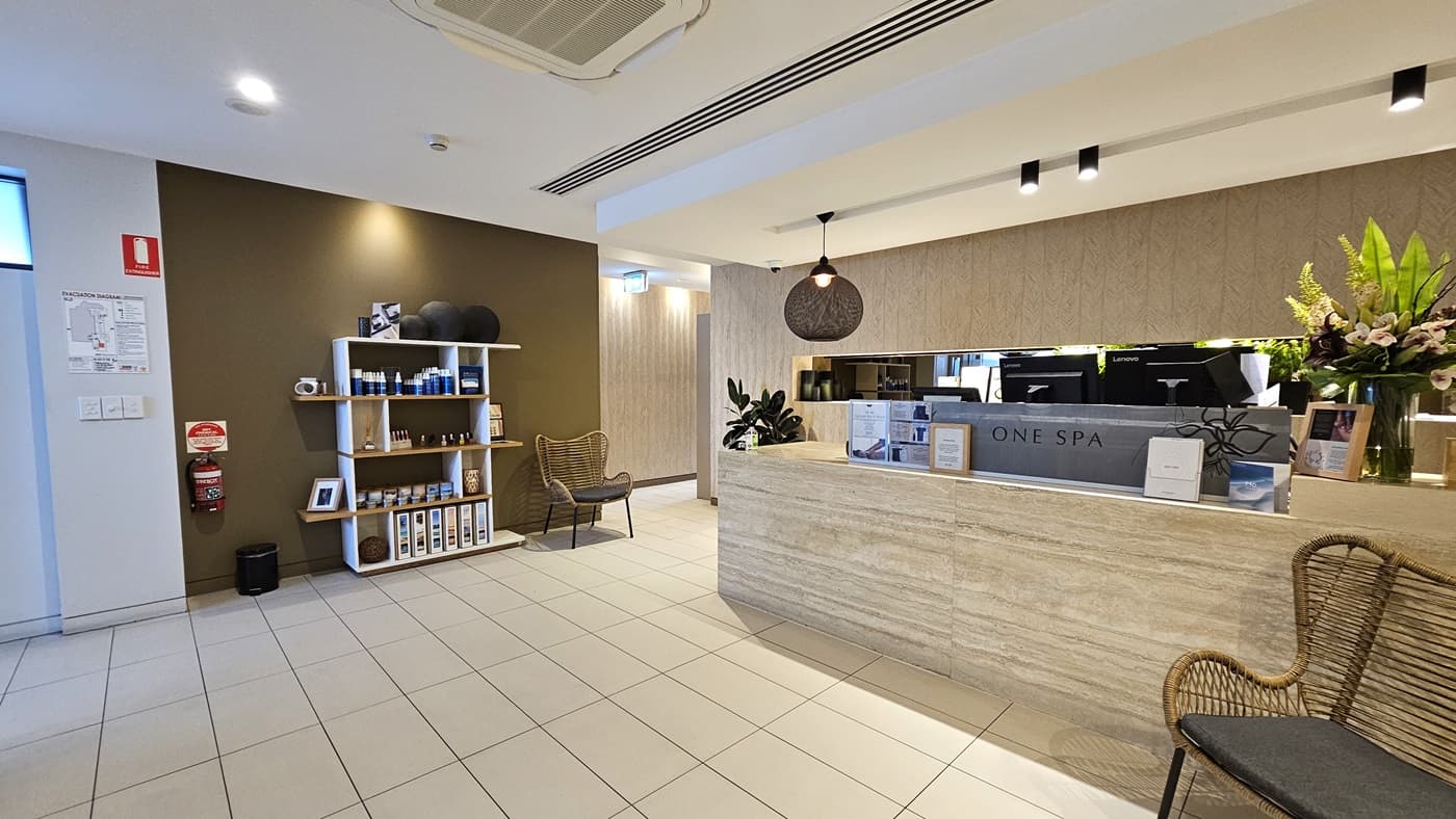 To the right is a reception desk with a timber-designed front, holding brochures. On the left, a display rack against a brown wall showcases items for sale. A rattan chair is on the right, and a rubbish bin and fire extinguisher are on the left. The wall features intricate design, with a white ceiling hosting cove lights and suspended stylish fixtures.