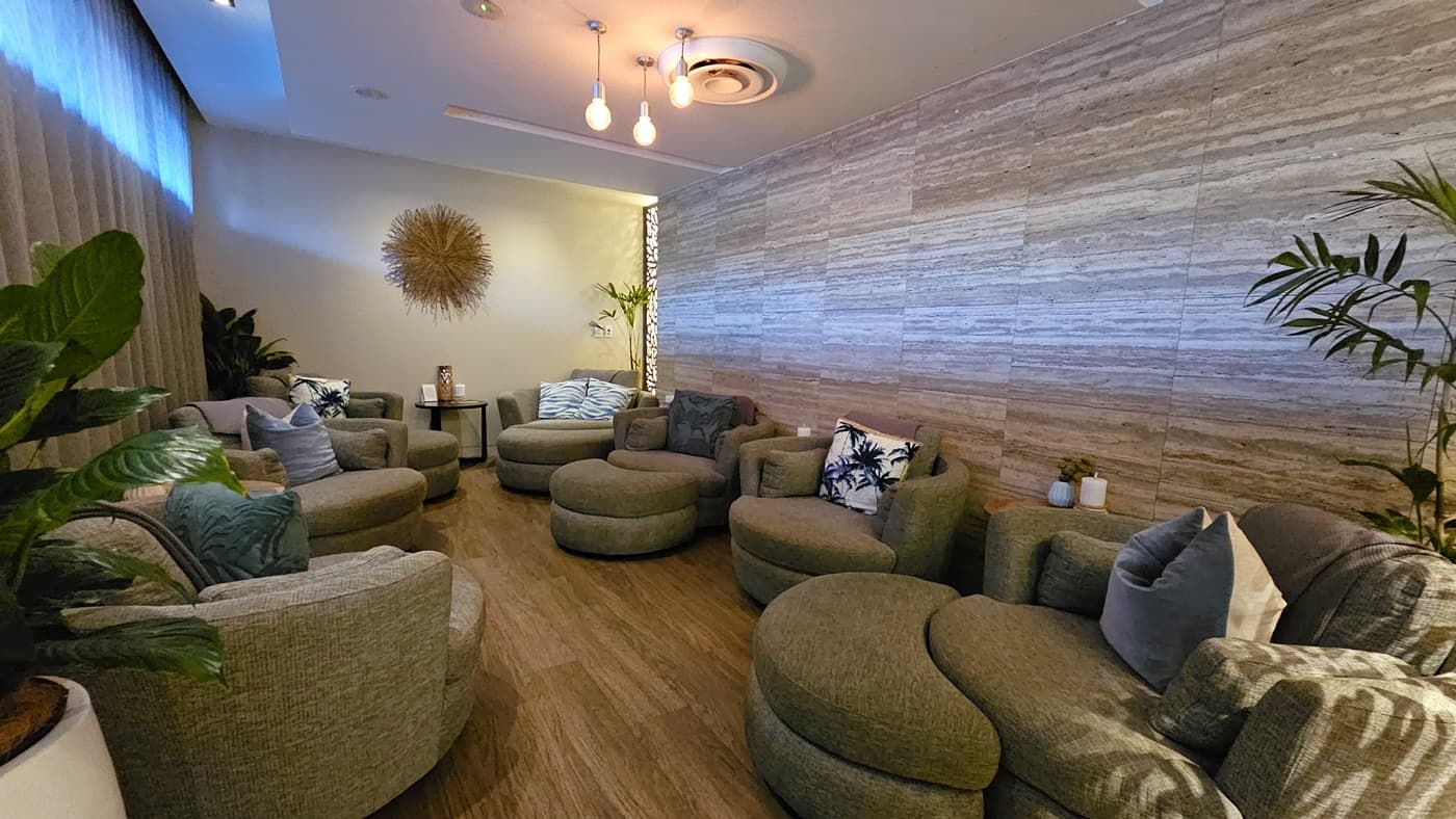 A room with grey cushioned chairs lined up on both sides. Large indoor plants are in each corner. The left wall has floor-to-ceiling grey curtains, the right wall features a timber design, and the middle white wall has a ball of thread decoration. The ceiling is white with light fixtures, and the floor has timber tiles.