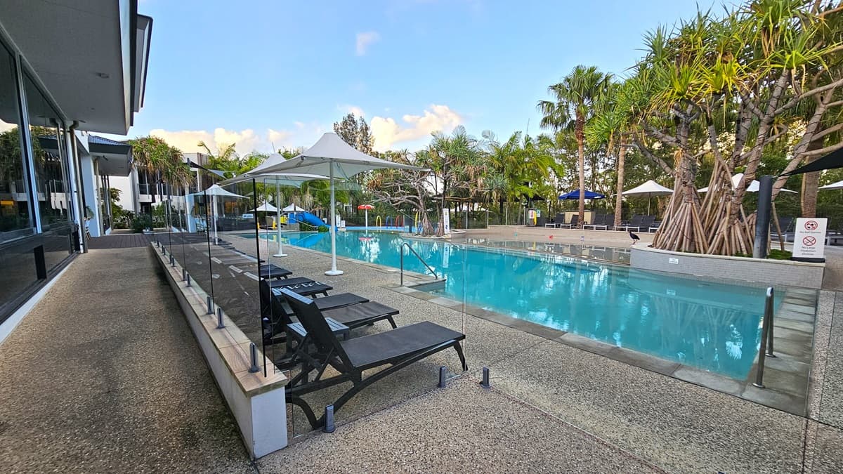 In the middle is a pool with handrails, surrounded by large white umbrella shades. Black sun lounge chairs are arranged on the left side of the pool deck. To the right is a concrete plant box with palm trees and light fixtures. A warning sign is placed below. At the far end, more sun lounge chairs are covered by large umbrella shades on the pool deck. Palm trees surround the pool area.