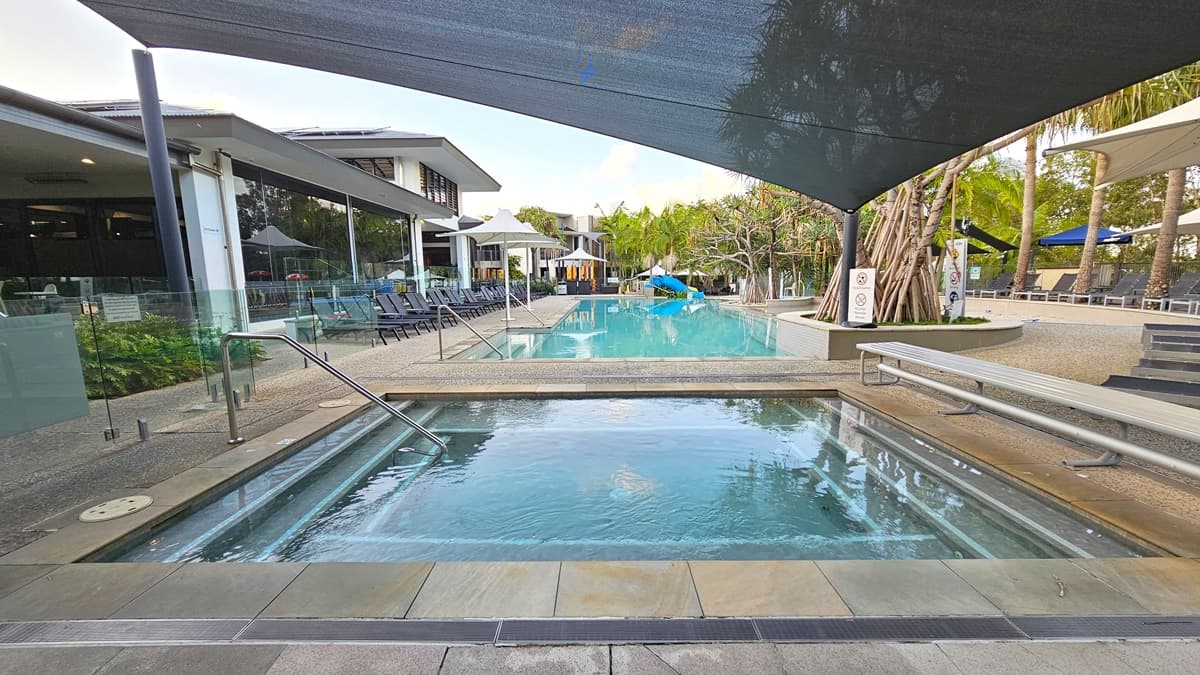 A pool with steps and handrails. Bench seating is on the right. A large black shade cloth covers the pool. Beside it is a bigger pool also with handrails. Sun lounge chairs are on the left deck. White umbrella shades are on both sides. Tall palm trees surround the area. The facility building is on the left.