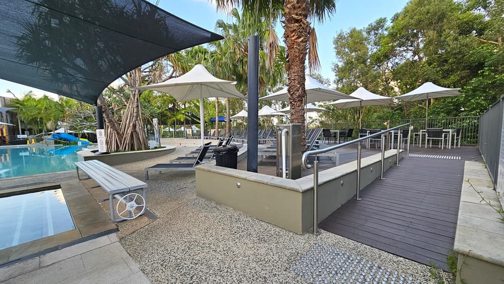 On the left is a pool shaded by a black shade cloth, with a larger pool behind it. Bench and sun lounge chairs are arranged on the right pool deck under large umbrellas. To the right, a wooden ramp with railings and truncated domes leads to an elevated pool deck section with tables and chairs under large white umbrella shades. A drinking fountain is positioned at the corner, next to a black rubbish bin. Tall palm trees and other trees surround the pool area.