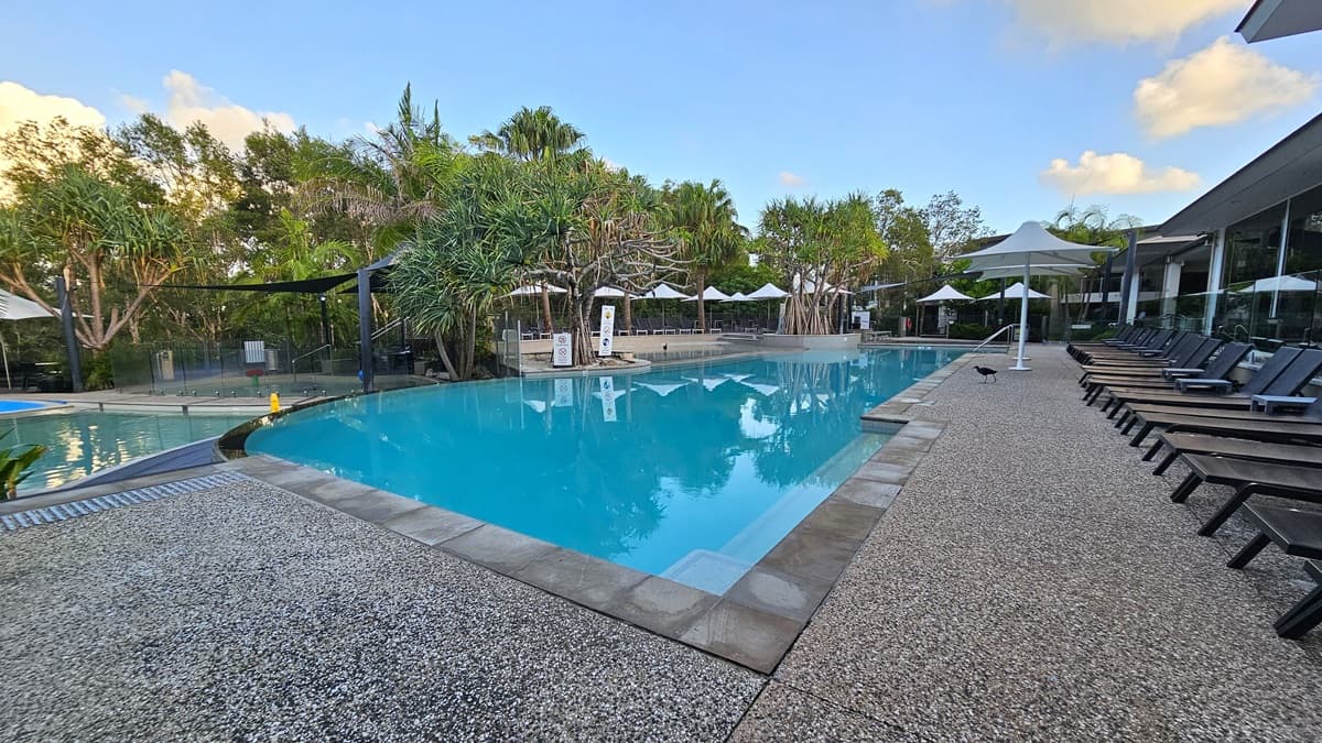 In the centre is a pool with hand rails and stepped entry, encircled by large white umbrellas. Black sun loungers line the pool deck on the right. On the left, a smaller pool is shaded by a cloth, with a nearby kids' pool. Various trees, including palm trees, adorn the pool area. Warning signs are visible.