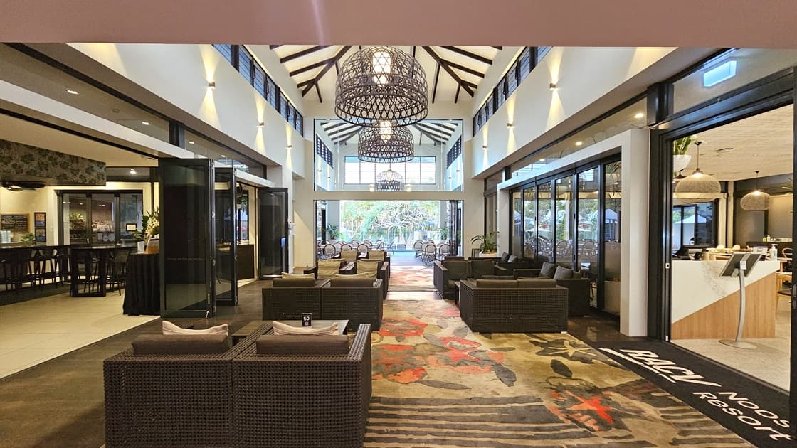 In the middle, there is arranged seating composed of cushioned chairs made of brown rattan and tables. To the right is the host station desk featuring a reception desk with a wooden design. Rows of arranged tables and chairs occupy both sides. The high ceiling features an intricate design with stylish light fixtures. The floor combines black and white tiles with colourful matting bearing the facility name.