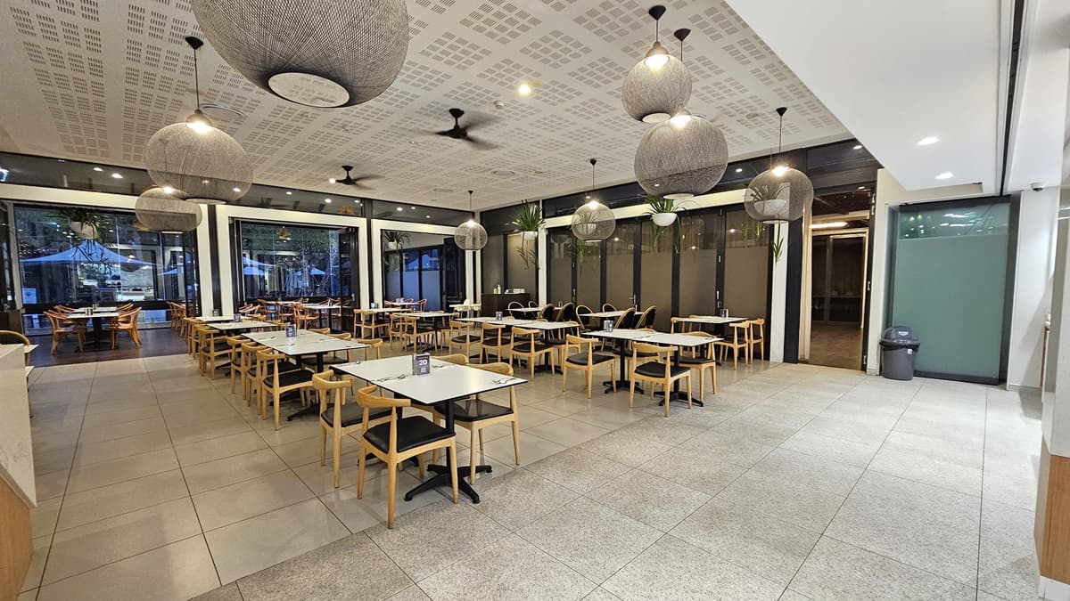 Movable chairs and tables neatly arranged in rows in an open space. Plain white ceiling with striking lighting fixtures resembling balls of thread suspended next to ceiling fans and potted plants. Dotted cream floor and wall tiles matched in colour. Glass partition runs along the left side. Glass door on the right leads to an additional room. A rubbish bin was placed nearby. Glass wall also borders the left section further inside the room.