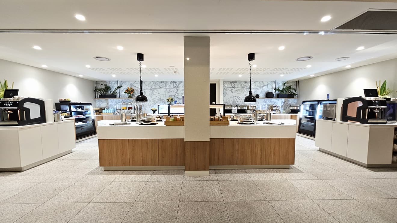 In the centre is a buffet section with food items displayed on the tables. Fridges on both sides showcase more food. Cream tiles cover the floor, matching the walls. The back wall has intricate plant decorations. The white ceiling has a slit design with LED lights and suspended light fixtures.