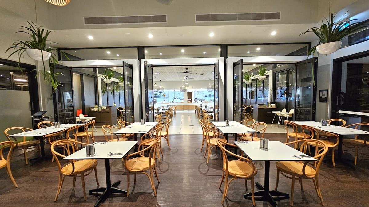 Movable chairs and tables are arranged in rows in the middle and on the sides, with utensils laid out. The white ceiling has a slit design with ceiling fans and stylish light fixtures resembling large thread balls hanging down. Ornamental plants also hang from the ceiling on both sides. The floor transitions from brown tiles to white. At the far end is the buffet section.