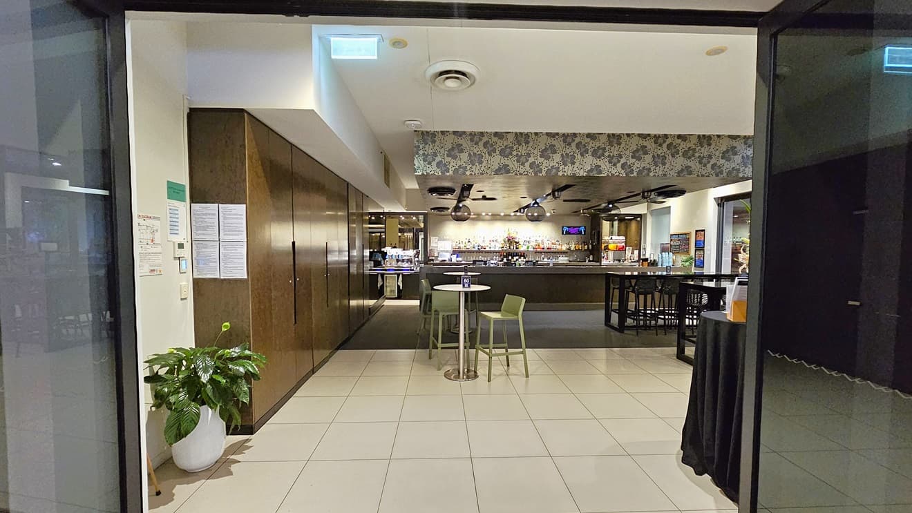 In the middle is the bar counter displaying wines, liquors, and glasses. Movable chairs and tables are positioned in front. The ceiling features an intricate floral design with stylish light fixtures and ceiling fans. The brown wall on the left features posters and reminders, with a pot of indoor plants below. The floor is tiled with white.
