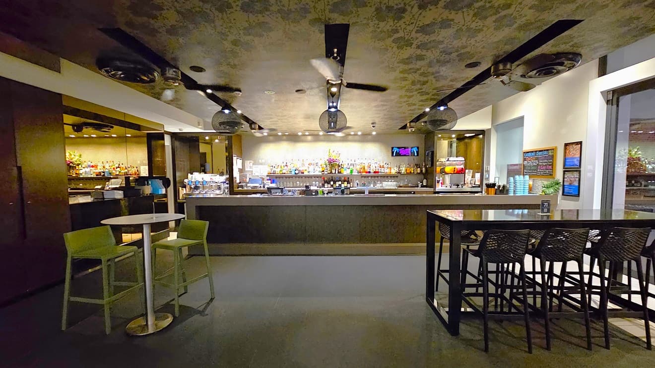 In the middle, there's a bar counter showcasing wines, liquors, and glasses. Movable chairs and tables are in front. The ceiling has an intricate floral design with stylish light fixtures and ceiling fans. The white wall on the right has colorful posters and reminders. The floor is tiled with white.