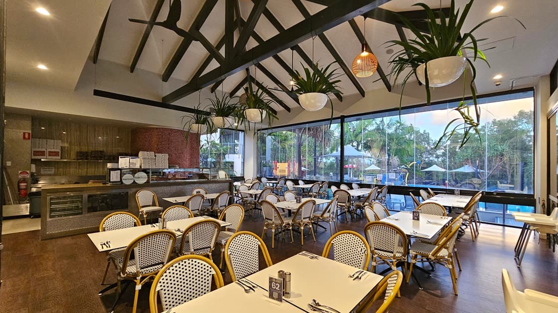 In the middle is a counter displaying food items. Movable chairs and tables are in front. The ceiling has a wooden design with stylish light fixtures; potted ornamental plants and ceiling fans hang from it. The white wall on the right has colorful posters and reminders. The floor is tiled with white. To the right, the glass wall offers a view of the pool outside.
