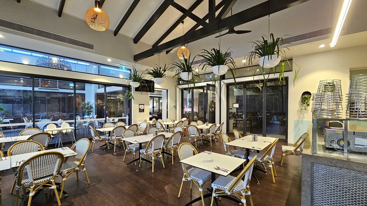 Tables and chairs are arranged. The ceiling has a wooden design with stylish light fixtures; potted ornamental plants and ceiling fans hang from it. The white wall on the right has colorful posters and reminders. The floor is tiled with white. To the left, more tables and chairs are arranged behind the glass wall. To the right is a counter table. The floor features polished timber.