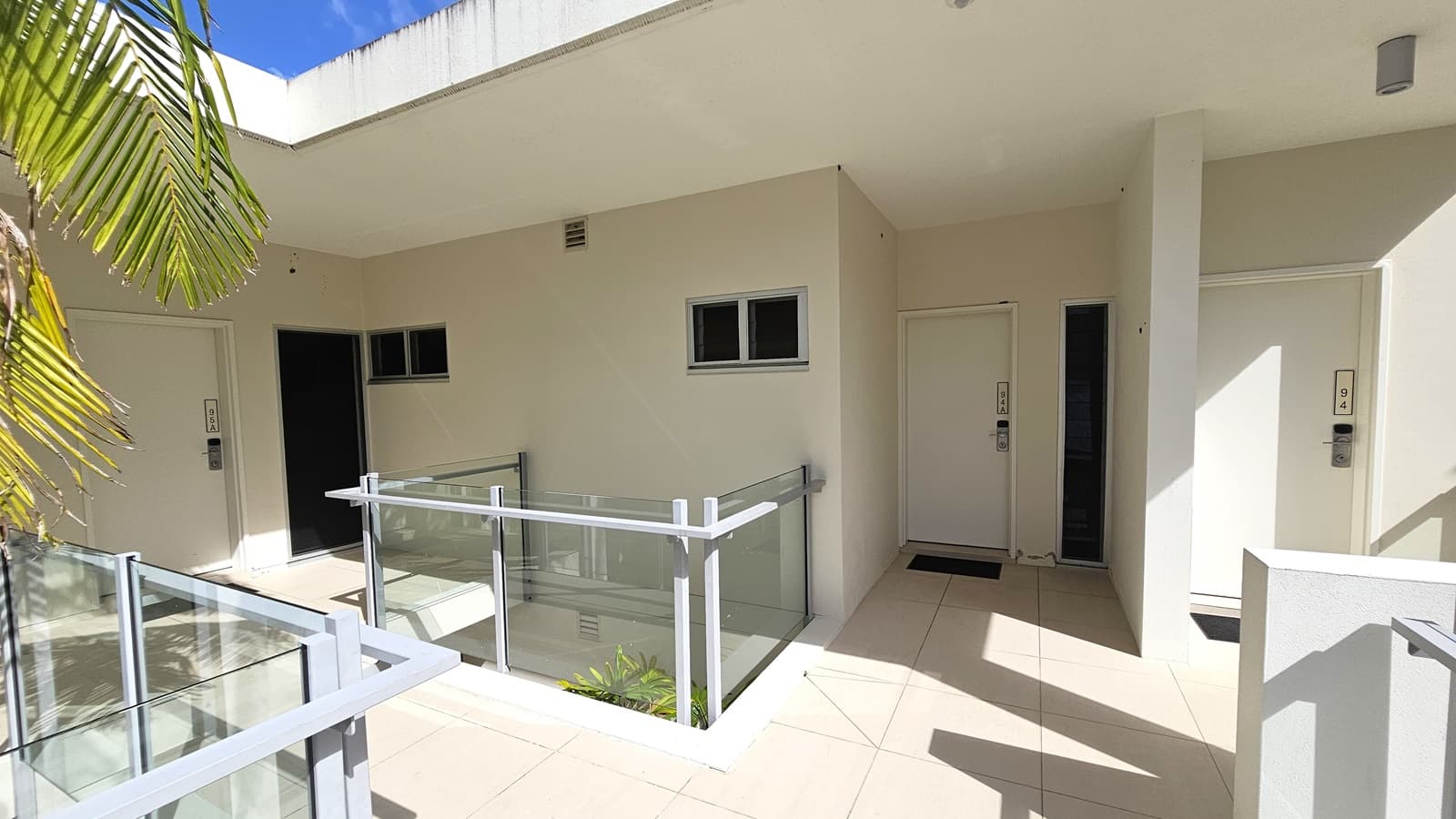 A staircase with handrails leads to the upper level, flanked by neatly trimmed shrubs and small trees. Tall palm trees are visible upstairs, with glass railings enclosing the upper level. The building walls are all white.