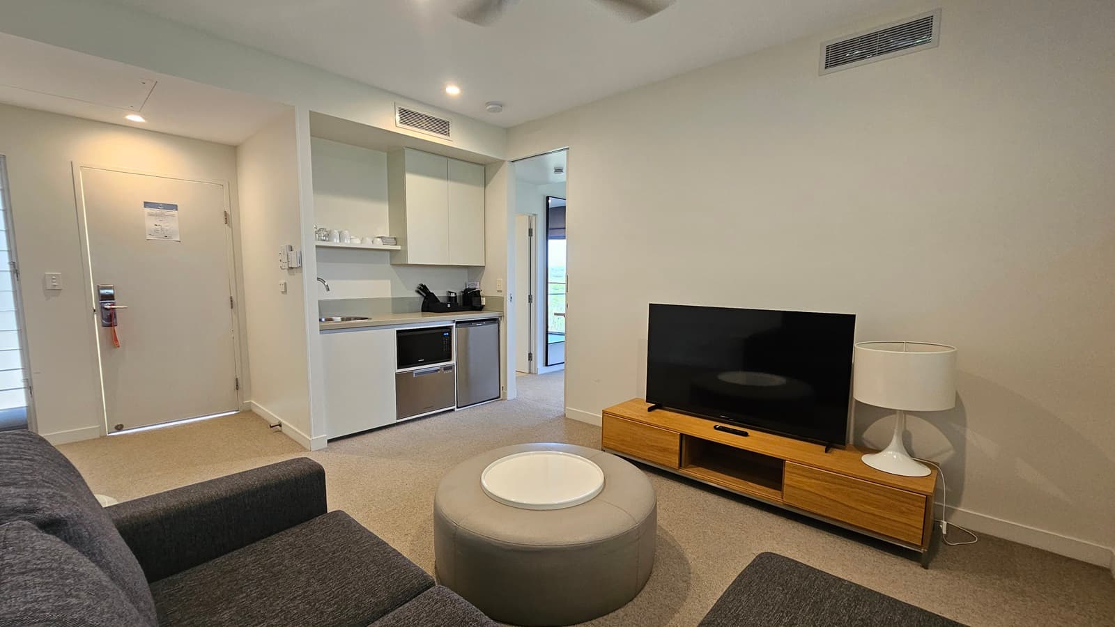 To the right, a flatscreen TV rests on a TV stand, accompanied by a lampshade. Adjacent is a lounge area with cushioned grey chairs and a round table. To the left is a cupboard area with a sink, followed by a door leading outside. The floor has grey carpet, while the wall and ceiling are white, lit by an LED light and ceiling fan.
