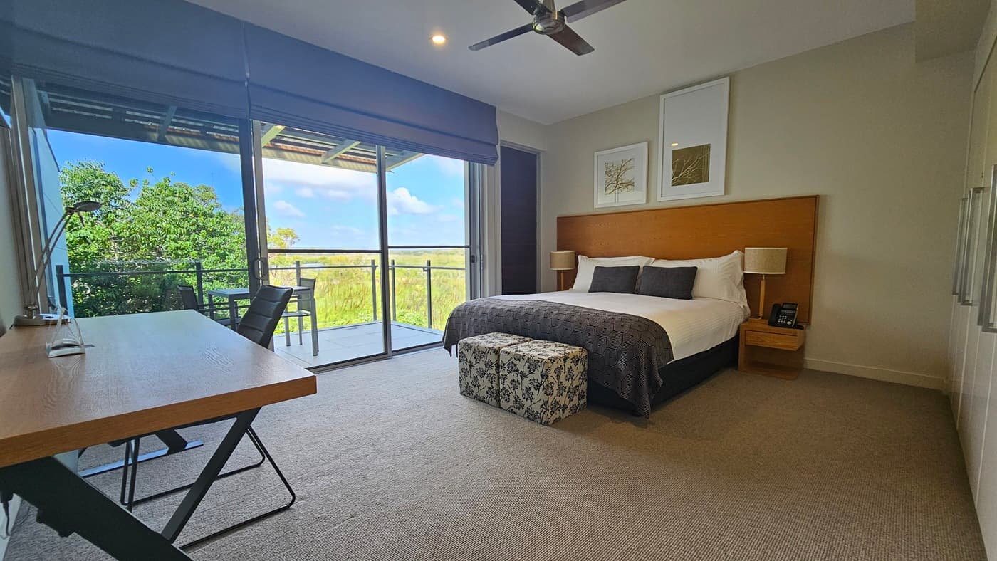 On the right, a king-size bed with a wooden headboard and matching bedside tables. Lampshades top the tables. A glass door to the balcony is on the left. Retractable curtains on the left wall. A wooden office table with a lamp is near the bed. Grey carpeted floor, white walls and ceiling with LED light and ceiling fan. Balcony has tables and chairs.