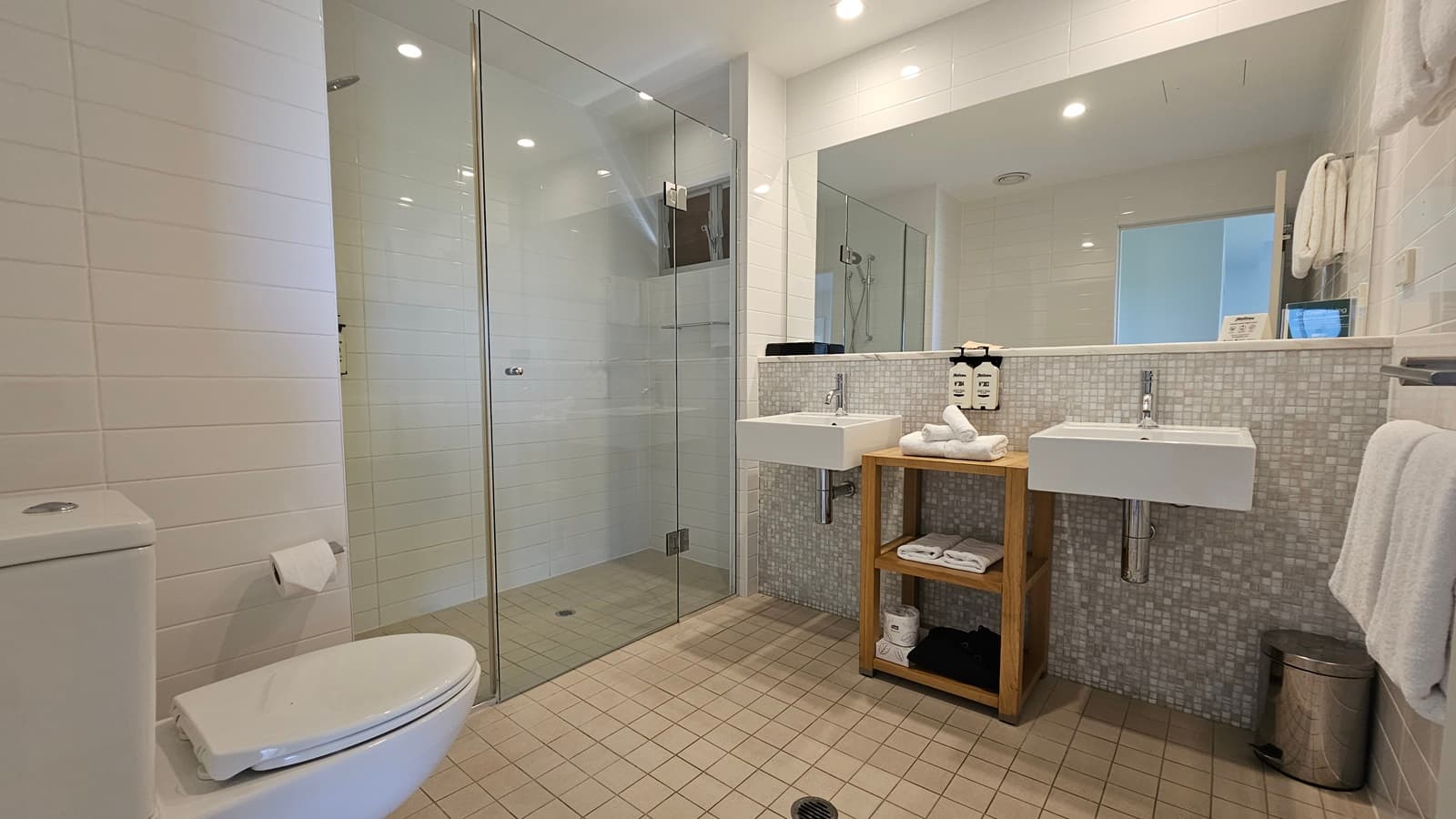 On the left, a toilet bowl with a tissue dispenser nearby. Next to it, a shower area enclosed by glass. To the right, a large mirror on a mosaic-design wall. Two hand basins mounted on the wall, with a wooden storage unit holding folded white towels between them. Liquid soap dispensers on the wall. A towel hangs from a holder to the right, along with a rubbish bin. Cream tiles cover the floor, and the ceiling is white with LED lights.
