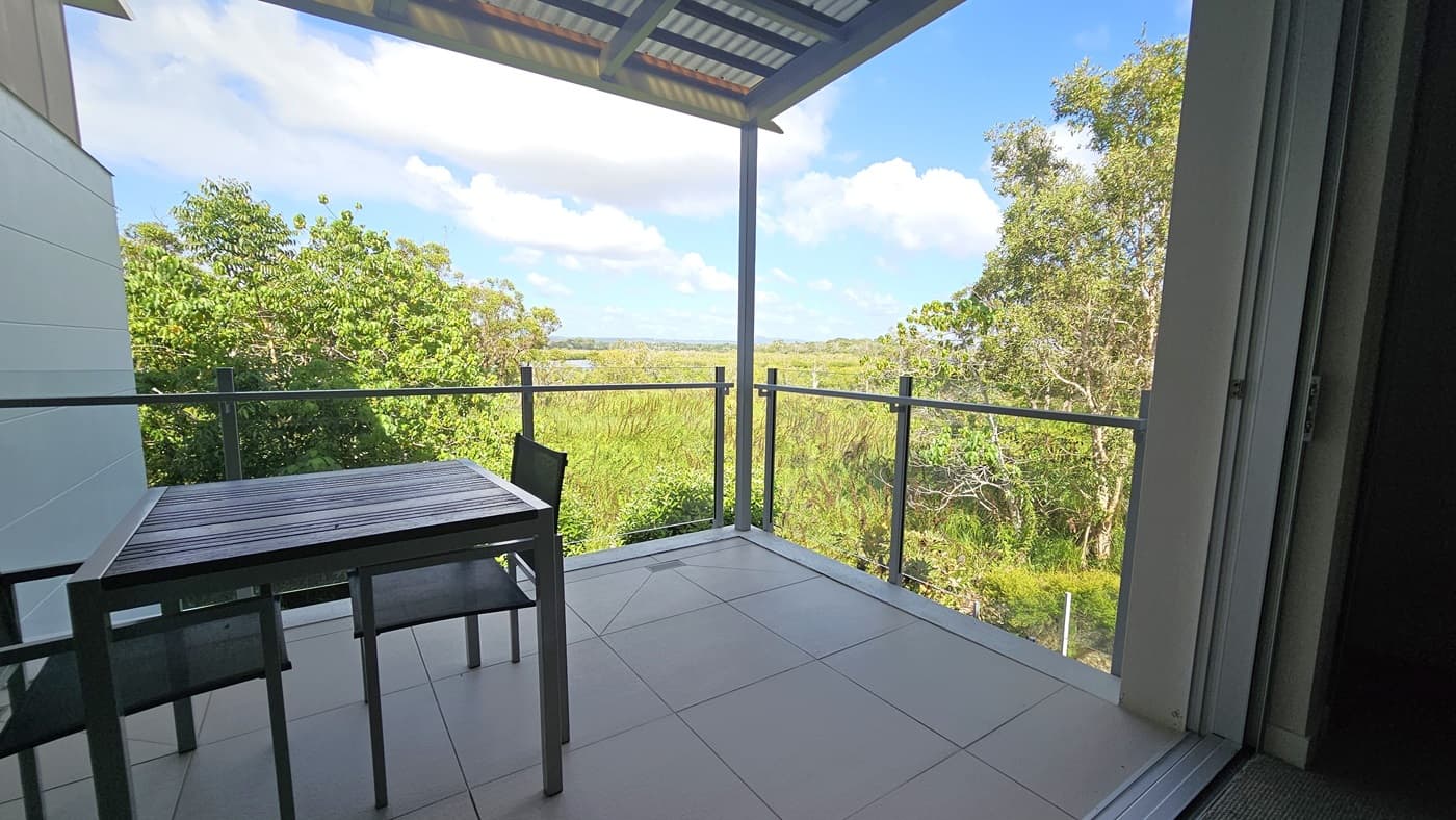 A serene view of lush green fields, grass, and tall trees. Movable table and chairs on the left. The balcony is enclosed by glass railings. The ceiling has corrugated roofing, while the walls and floor are tiled in white.