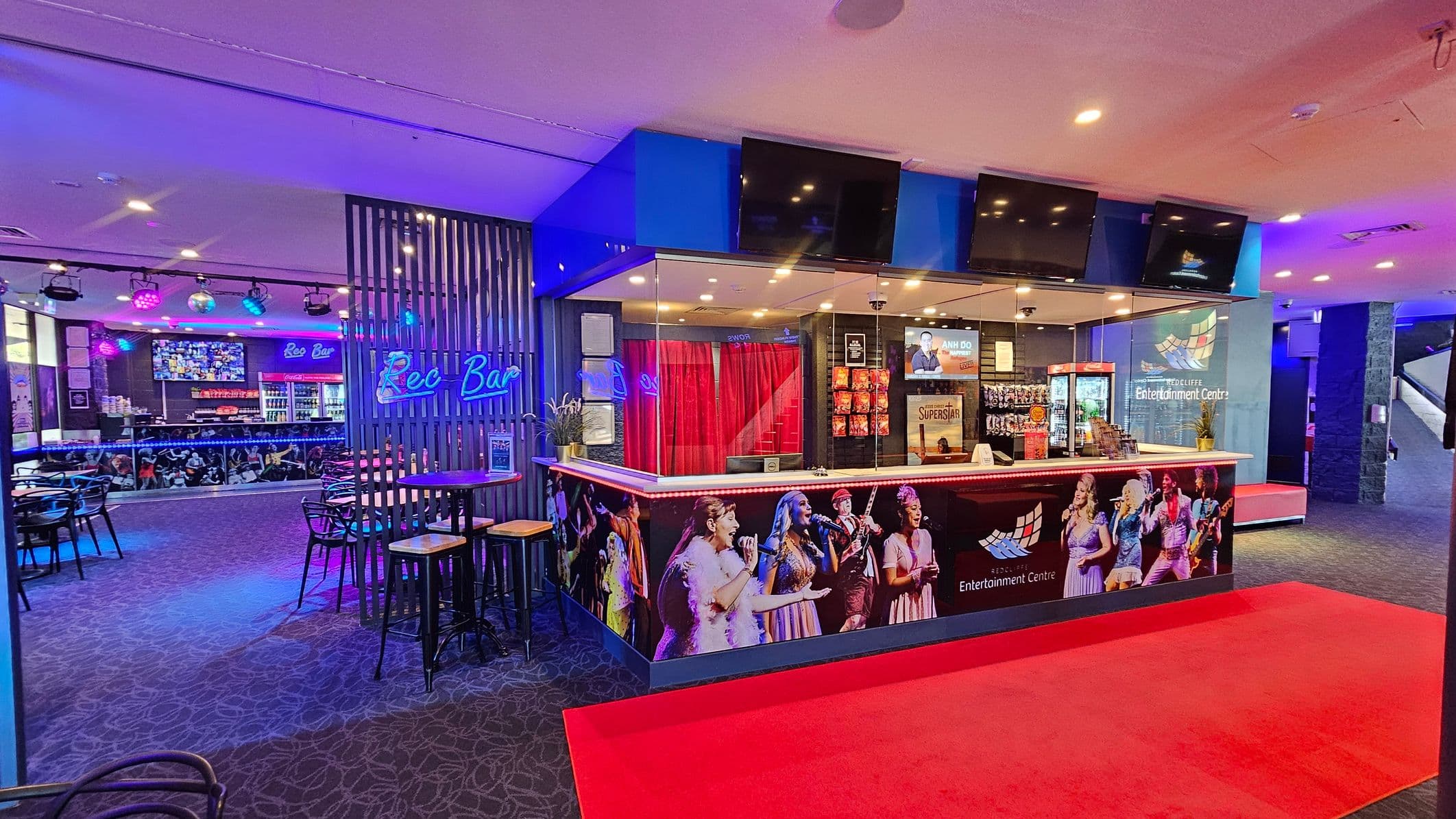 A vibrant entertainment venue with a bar area on the left, featuring a neon "Rec Bar" sign and several tables with stools. The bar counter displays various drinks and a large screen above. In the centre, a reception area with a counter adorned with images of performers. Behind the counter, red curtains and promotional posters are visible. The floor is carpeted with a red carpet leading to the counter, and the ceiling has recessed lighting.