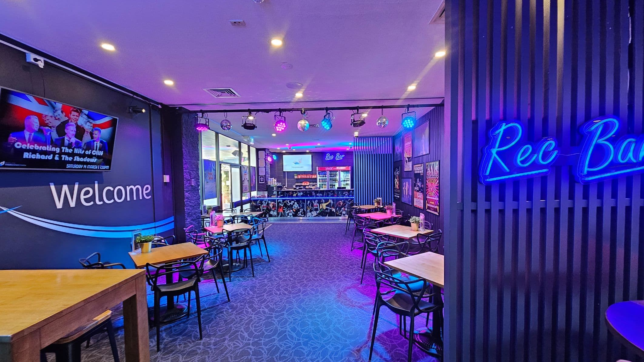A vibrant bar interior with a purple and blue lighting theme. On the left, a wall-mounted screen displays an event poster above the word "Welcome." Several wooden tables with black chairs are arranged throughout the space. On the right, a neon sign reads "Rec Bar" on a slatted wall. The bar area at the back features a counter with drinks and a screen above. The ceiling has spotlights and disco balls, and the carpet has a subtle pattern.
