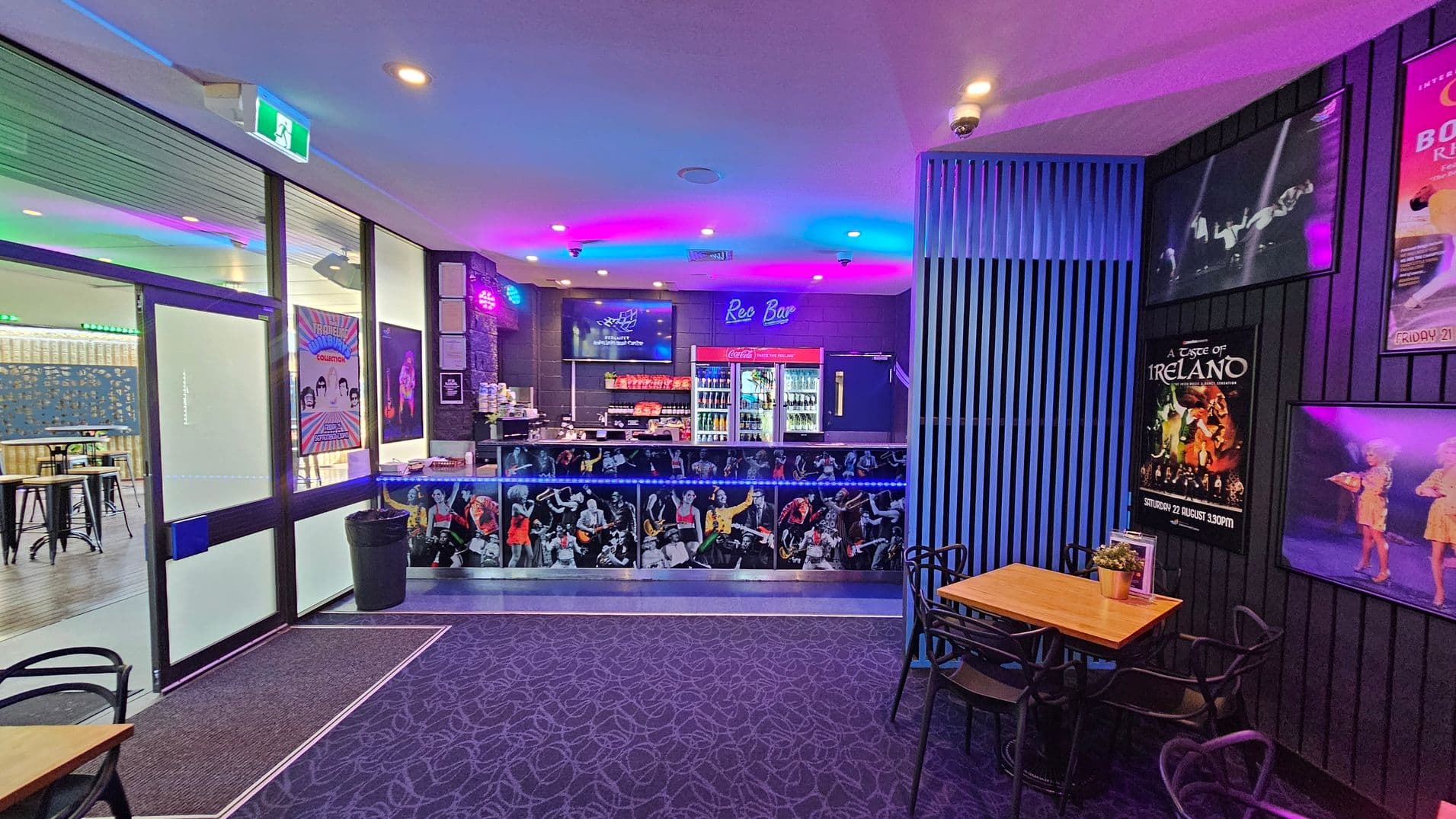 A bar area with a vibrant, colourful atmosphere. On the left, there are glass doors with posters and a glimpse of high tables and stools in the adjacent room. The centre features a bar counter decorated with images of musicians, with a neon "Rec Bar" sign above. Behind the counter, there are shelves with drinks and snacks. On the right, a small wooden table with black chairs is set against a wall adorned with various event posters. The ceiling has recessed lighting, and the floor is carpeted with a patterned design.