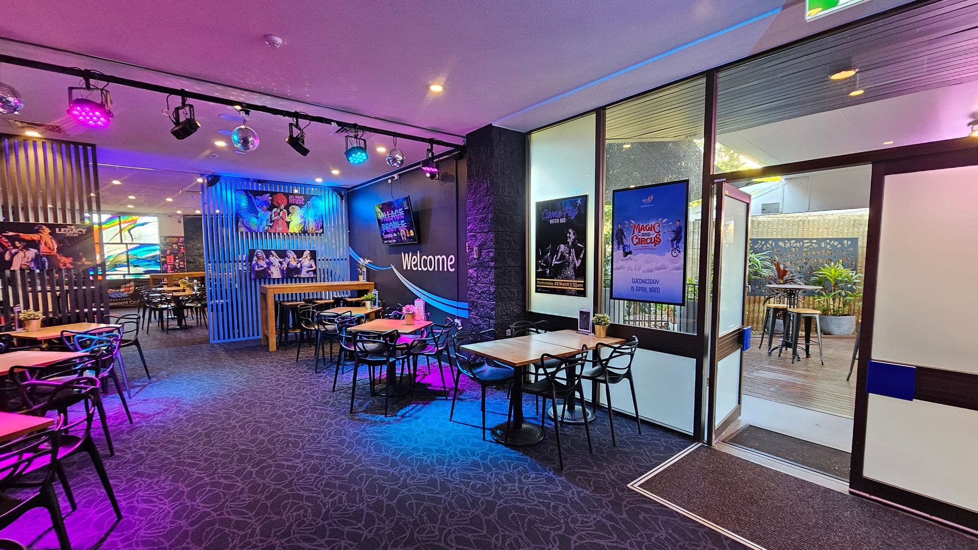 A vibrant indoor space with a patterned carpet floor and a ceiling featuring colourful spotlights and disco balls. On the left, there are tables and chairs, with posters on the walls. In the centre, a "Welcome" sign is visible on a dark wall. To the right, large windows and a glass door lead to an outdoor area with plants and high tables. Posters are displayed on the walls near the entrance.
