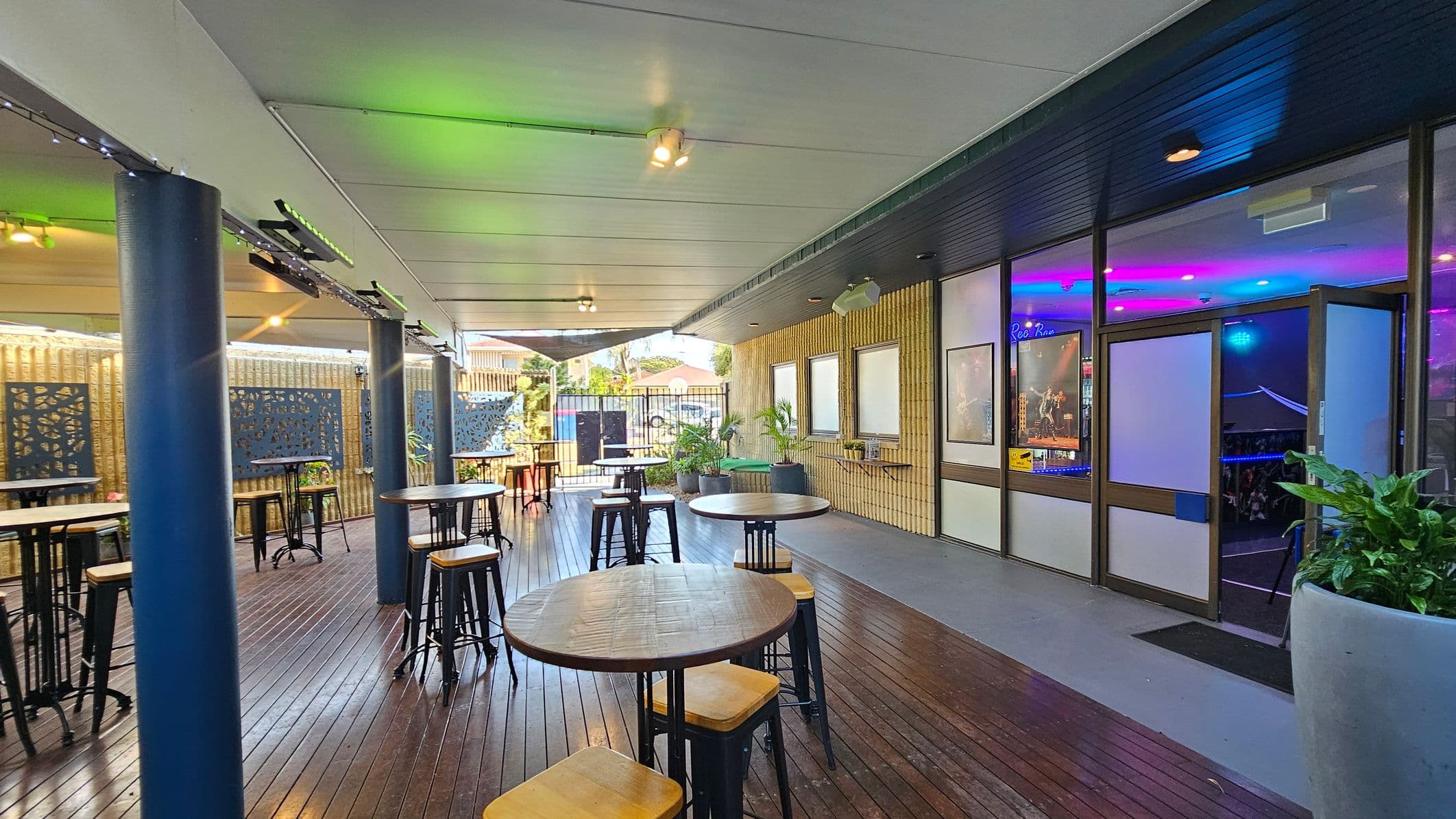 A covered outdoor area with wooden flooring and a white ceiling. On the left, there are several round wooden tables with high stools, and blue decorative panels on the walls. Blue columns support the ceiling. On the right, a glass door leads inside, where colourful lights are visible. Potted plants are placed near the entrance. The background shows a fence and some buildings.