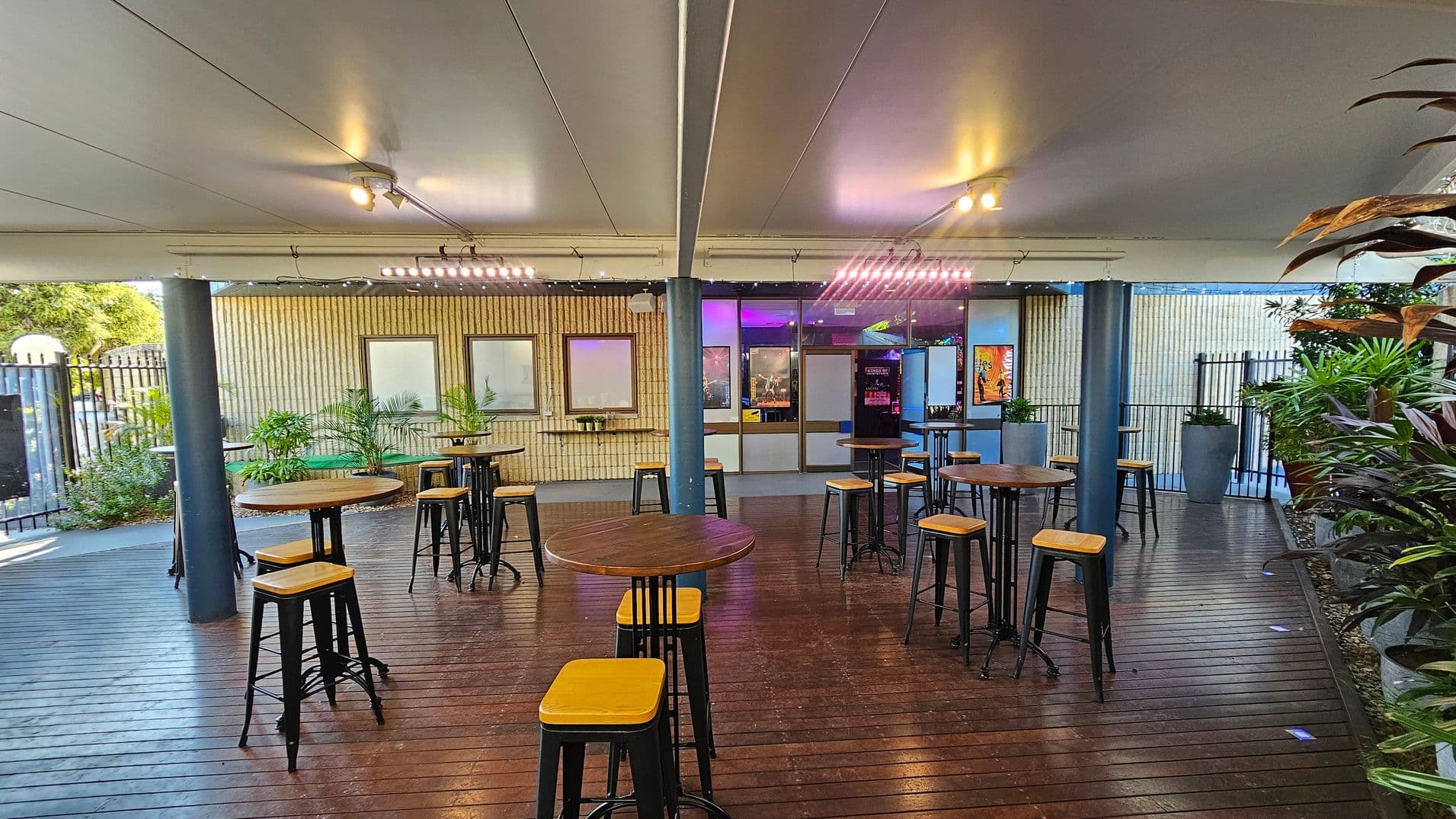 A covered outdoor seating area with wooden flooring and a white ceiling. In the centre, there are several round wooden tables with tall stools. The background features a wall with windows and a door, illuminated by purple and yellow lights. Potted plants are placed along the wall and on the right side. The left side shows a metal fence and more greenery. Ceiling lights provide additional illumination.