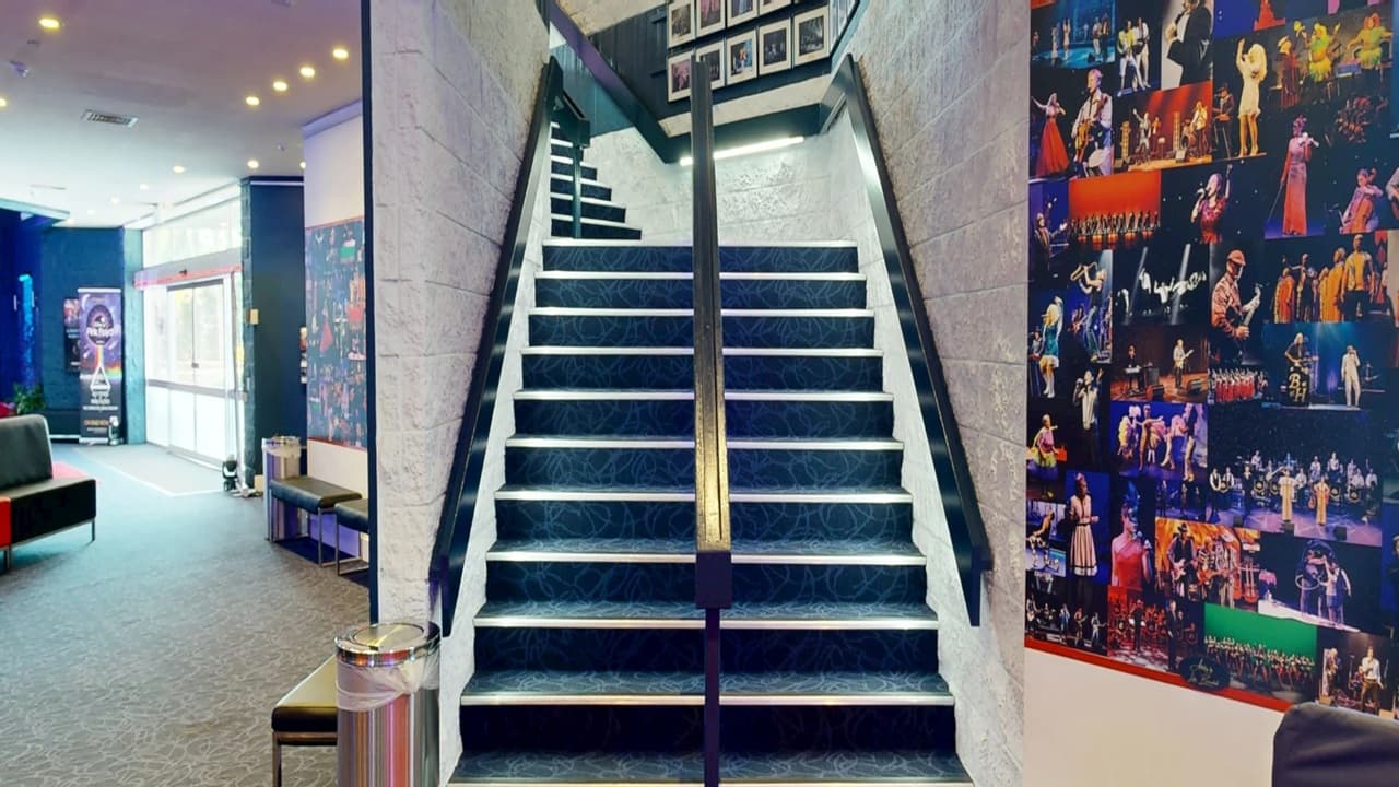 A staircase with dark blue carpet and white walls, featuring a central handrail, leads upwards. On the right, a wall is covered with vibrant performance photos. To the left, a seating area with a dark sofa and a small table is visible. The floor is carpeted in a similar dark tone. The ceiling has recessed lighting, and a large windowed entrance is in the background, allowing natural light to enter.