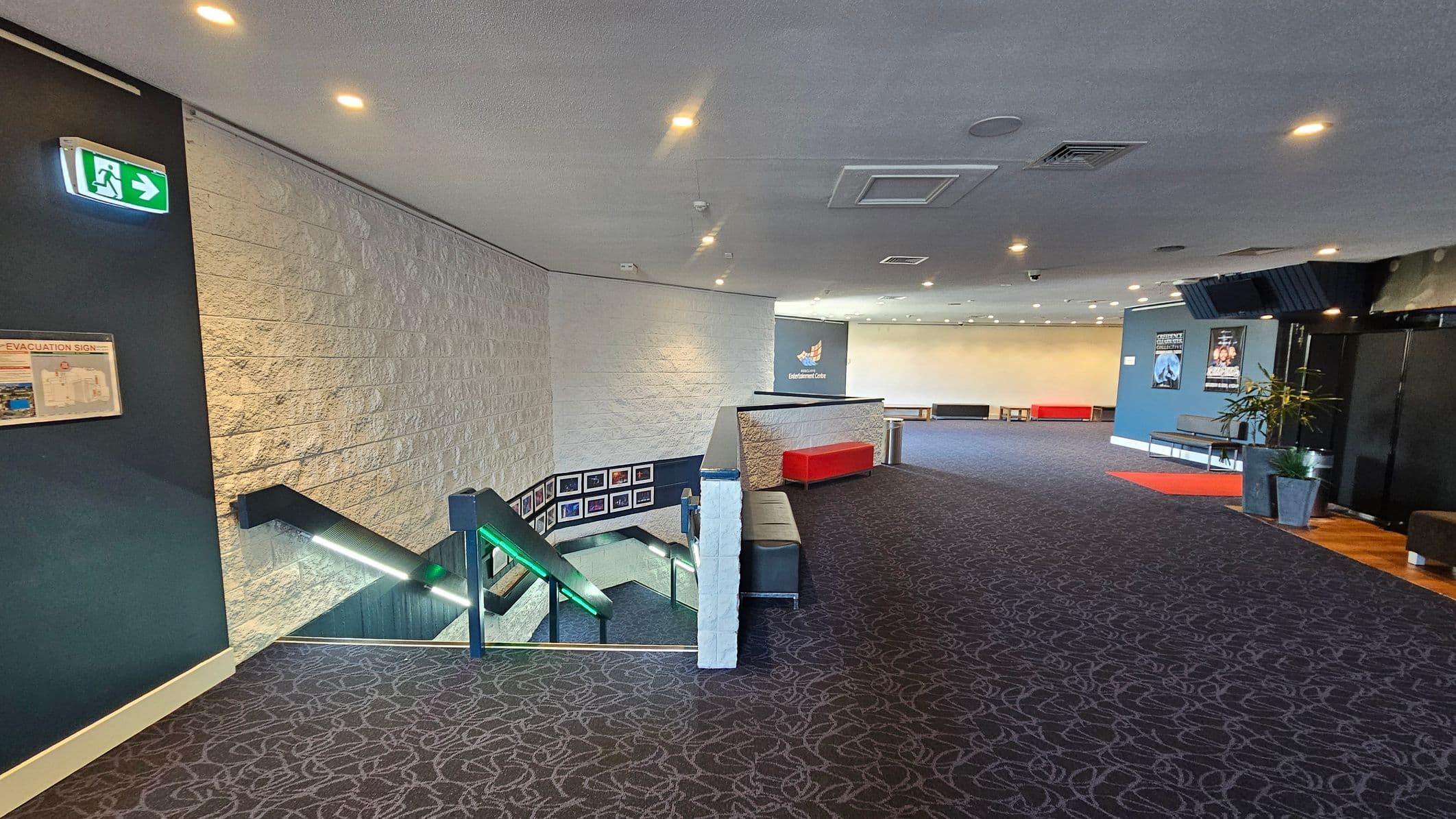 A spacious interior with a patterned carpeted floor. On the left, a staircase with illuminated handrails descends, and a green exit sign is visible above. The wall is textured with a light colour. In the centre, a red bench sits against a low wall. The right side features a blue wall with posters and a potted plant. The ceiling is fitted with recessed lighting and air vents.