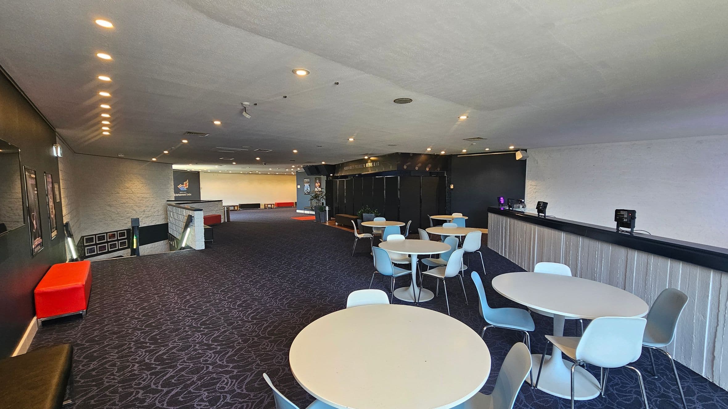 A spacious room with a dark patterned carpet and a white ceiling with recessed lighting. On the right, there is a long bar counter with electronic devices on top. In the centre, several round tables with light blue and white chairs are arranged. On the left, a red bench sits near a staircase leading downwards, with framed pictures on the wall. The back wall features a sign and more seating.