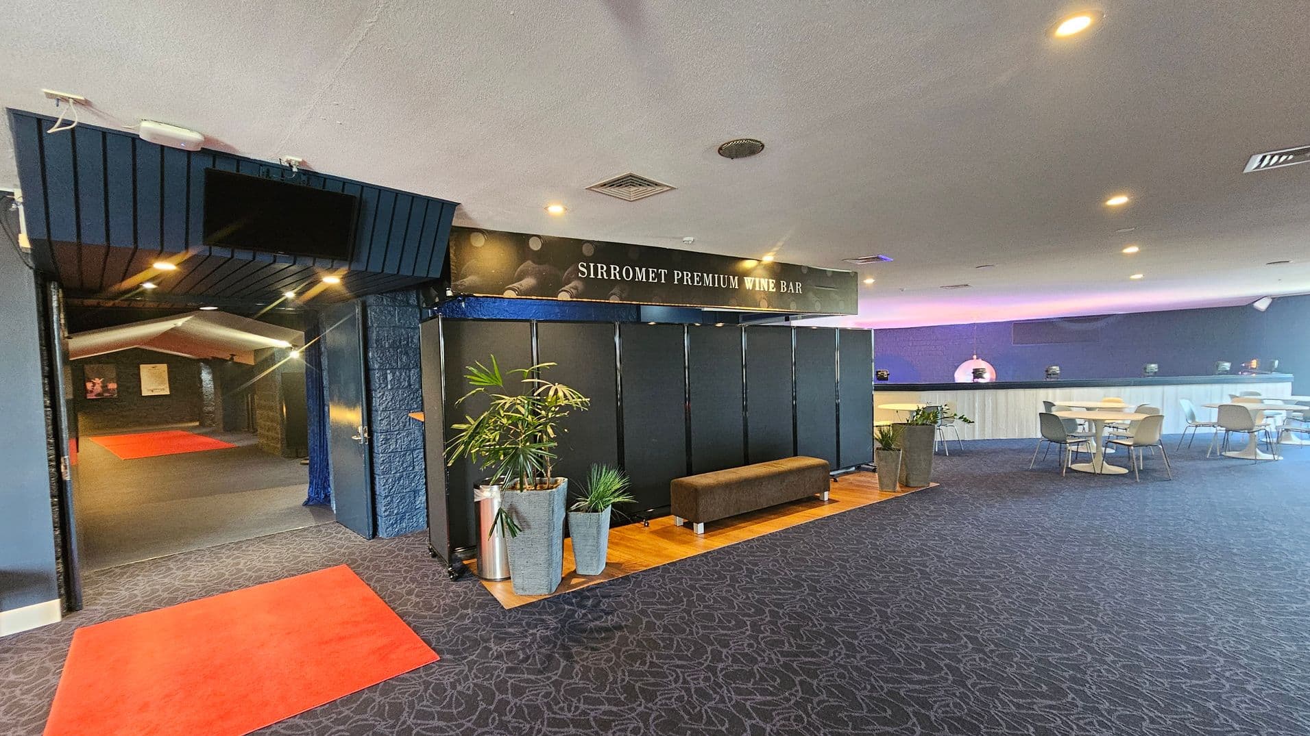 Entrance to a wine bar with a sign reading "Sirromet Premium Wine Bar" above. On the left, a hallway with red carpets leads further inside. In the centre, a seating area with a brown bench and potted plants on a wooden floor. On the right, a spacious area with tables and chairs, and a long counter against the wall. The ceiling is white with recessed lighting, and the floor is covered with patterned carpet.