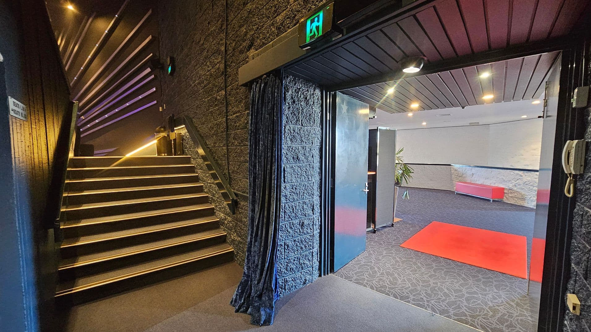 On the left, a staircase with illuminated steps ascends alongside a textured wall. Above the stairs, a series of horizontal lights create a pattern. On the right, an open door leads to a room with a red carpet on a patterned floor. The room has a red bench against a light-coloured wall and a potted plant. The ceiling is dark with recessed lighting. An emergency exit sign is above the door. A curtain hangs beside the door.