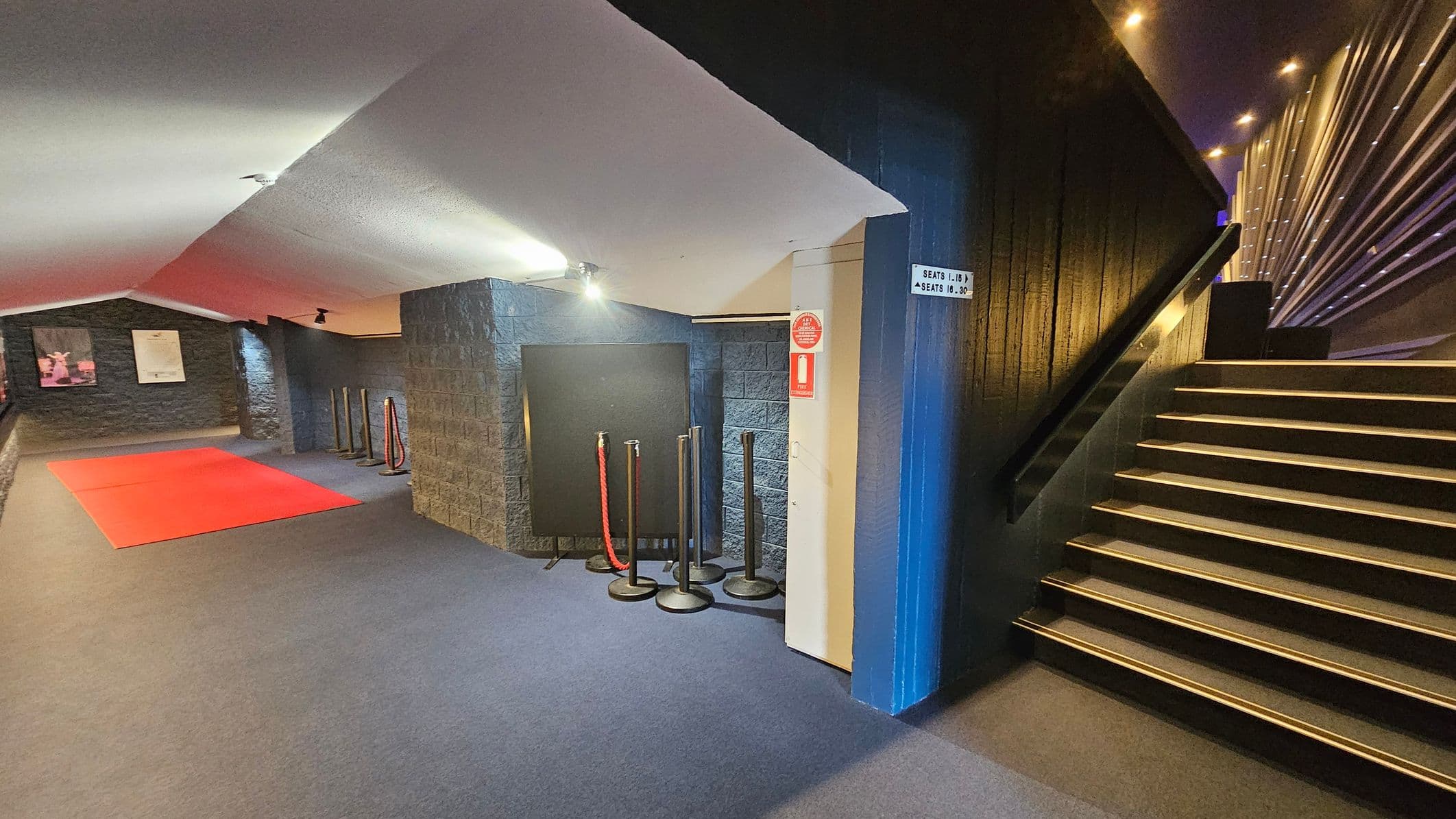 A dimly lit interior with a red carpet on the left side of the floor, leading towards a dark stone wall with two framed posters. In the centre, there are several stanchions with red ropes. On the right, a staircase with illuminated steps ascends alongside a textured wall with vertical lines. The ceiling is slanted and white, with spotlights providing soft lighting. A sign on the wall indicates seating directions.