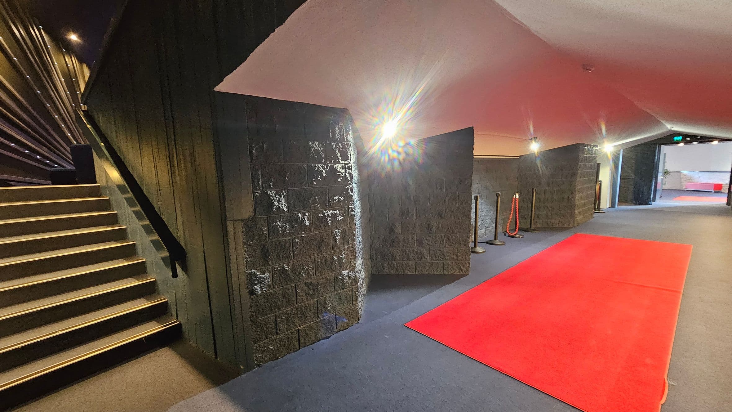 A hallway with a red carpet on the right side, leading towards an open area in the distance. The floor is covered with a dark carpet. On the left, there is a staircase with golden-edged steps and a black handrail. The walls are textured and dark, with bright lights illuminating the space. The ceiling is angled and light-coloured. There are stanchions with red ropes near the walls.