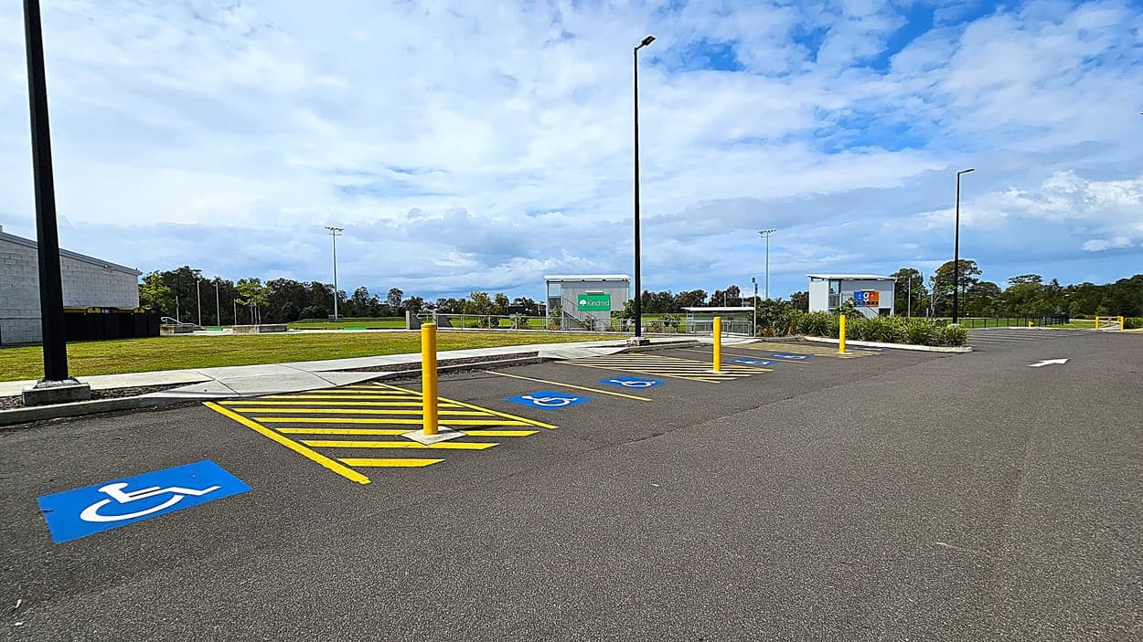 On the left, there is a grey building with a flat roof and large windows, alongside a smooth concrete pathway. To the right, several designated parking spaces are marked with blue symbols for disabled access, and yellow striped areas indicate no parking. In the background, there are light poles and a green sign, with a grassy area beyond. The asphalt surface of the parking lot is well-maintained, and the overall layout is spacious, allowing for easy navigation.