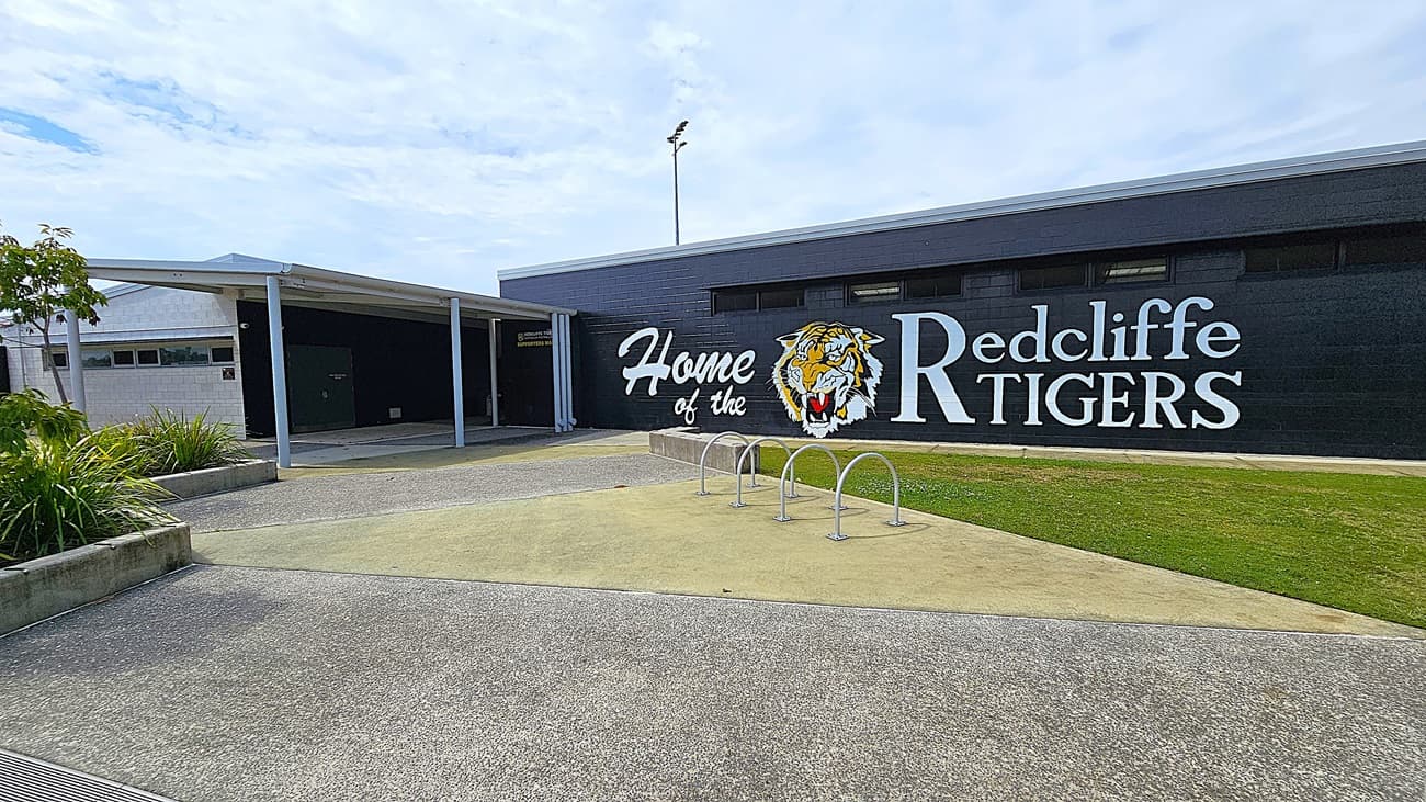 On the left, there is a white building with a sloped roof and a covered walkway leading to a dark door. Adjacent to it, a large black wall features the text "Home of the Redcliffe Tigers" alongside a tiger graphic. To the right, the wall continues with several windows. In the foreground, a smooth concrete area is visible, with a bike rack and patches of grass.