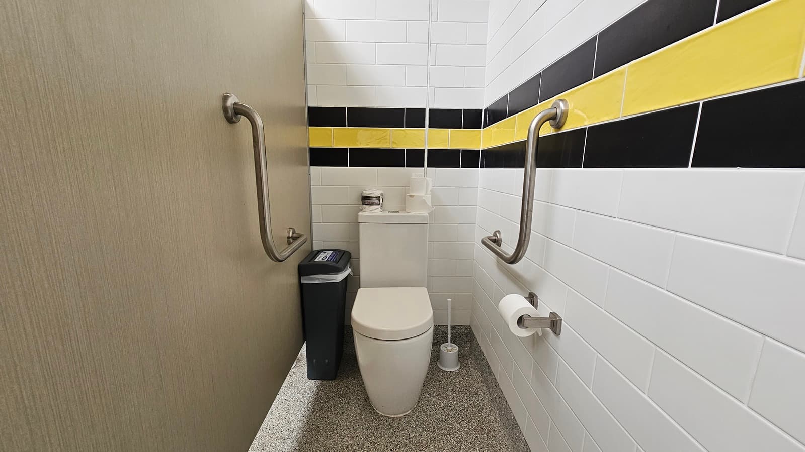 On the left, a light wooden door leads into the cubicle, with a smooth surface and a small handle. The walls are tiled with white tiles, accented by horizontal black and yellow stripes. In the centre, a modern white toilet is positioned against the wall, with a toilet roll holder beside it. A black bin is located to the left of the toilet. On the right, stainless steel grab bars are mounted on the wall for support.
