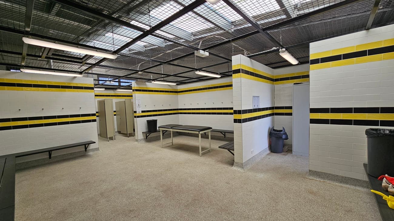 On the left, a light wooden door with a handle leads into the cubicle, alongside a stainless steel grab bar mounted on the wall. In the centre, a white toilet is positioned against the wall, with a toilet roll holder to the right. A black bin is located next to the toilet. The walls are tiled with white tiles, featuring horizontal black and yellow stripes.