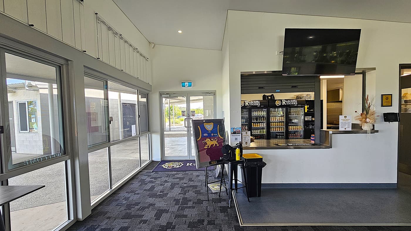 On the left, there are large glass doors leading outside, with a welcome mat featuring a logo. A framed jersey hangs on a stand nearby. To the right, a service counter is visible, with a display of drinks in glass-fronted refrigerators and a television mounted above. The floor is covered in dark carpet with a subtle pattern, while the ceiling is flat and features recessed lighting.