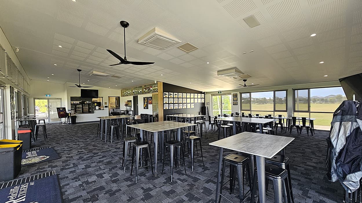 On the left, there are large glass doors leading outside, with a welcome mat in front. A bar area is visible, featuring shelves with drinks and a television mounted above. To the right, several tables with metal stools are arranged, and framed pictures hang on the wall. The floor is covered in dark patterned carpet, while the ceiling is flat with recessed lighting and ceiling fans.