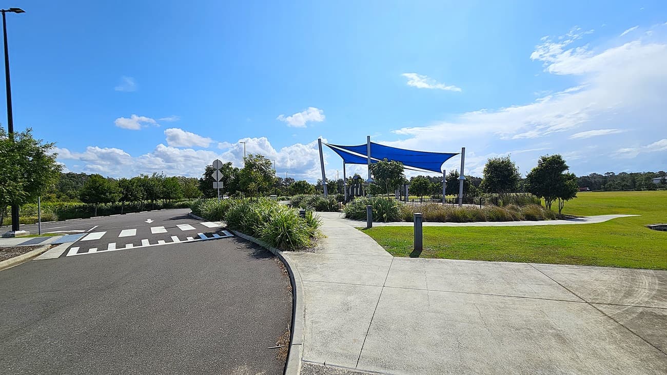 On the left, there are two large blue shade sails supported by tall silver poles, providing cover over a play area. Beneath the sails, a playground structure features a slide and climbing elements, surrounded by a soft, tan-coloured ground cover. To the right, a pathway leads towards a grassy field, with several trees in the background. The floor is made of smooth concrete, and the area is bordered by low fencing.