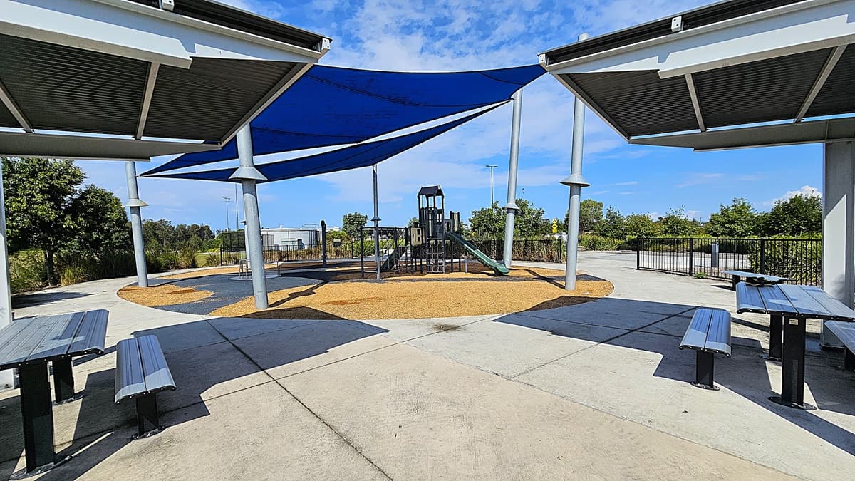 On the left, there are two metal picnic tables with attached benches, positioned on a smooth concrete surface. A blue shade sail is supported by silver poles above the tables. In the centre, a playground structure features a slide and climbing elements, surrounded by a soft, tan-coloured ground cover. To the right, the pathway curves away, leading to a fenced area with greenery. The floor is primarily concrete, and the ceiling consists of the shade sail overhead.