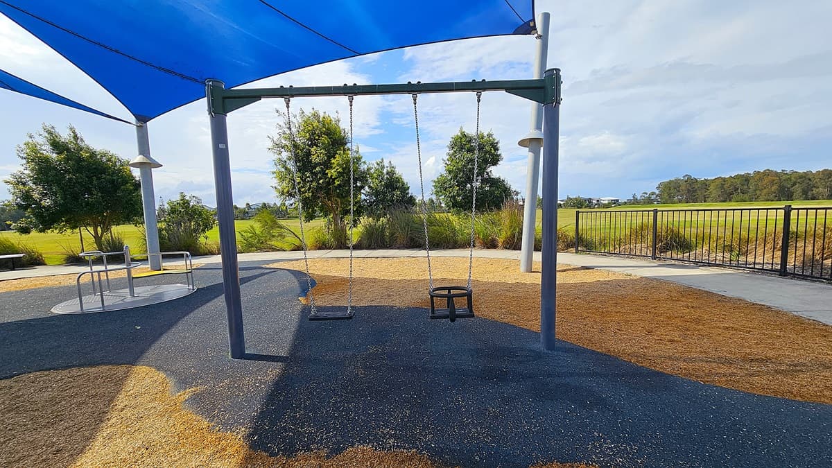 On the left, there is a swing set with two swings, one designed for a child and the other a standard swing, both hanging from a sturdy frame. The ground beneath the swings is covered in a mix of dark rubber matting and tan-coloured soft material. To the right, a circular metal table with attached seating is visible, surrounded by low shrubs and grass. The area is shaded by a large blue shade sail supported by tall poles. The floor is primarily a combination of rubber and natural ground cover.