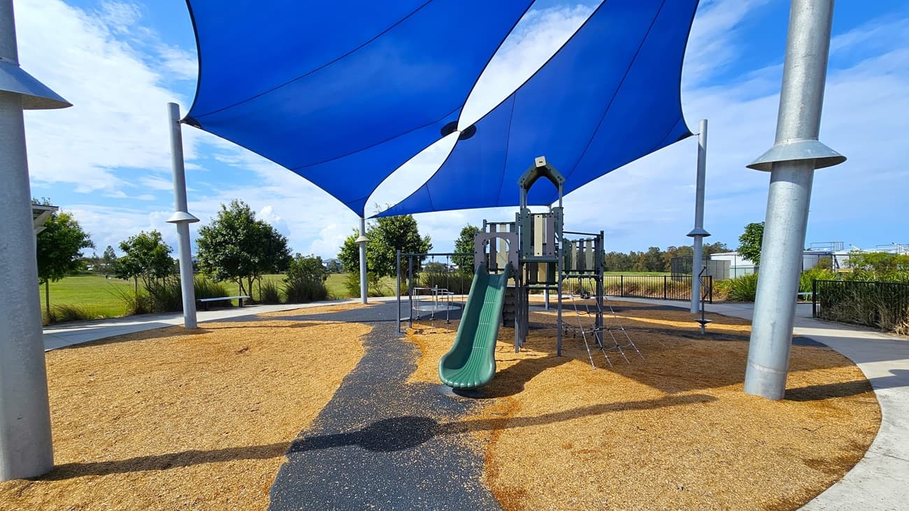 On the left, a green slide is attached to a multi-level playground structure, which includes climbing features and a small platform. The ground is covered with tan wood chips, providing a soft surface for play. To the right, there are several metal poles supporting a large blue shade sail that covers the play area. In the background, there are low shrubs and trees, with a paved pathway visible. The floor is primarily wood chips, and the ceiling consists of the shade sail.